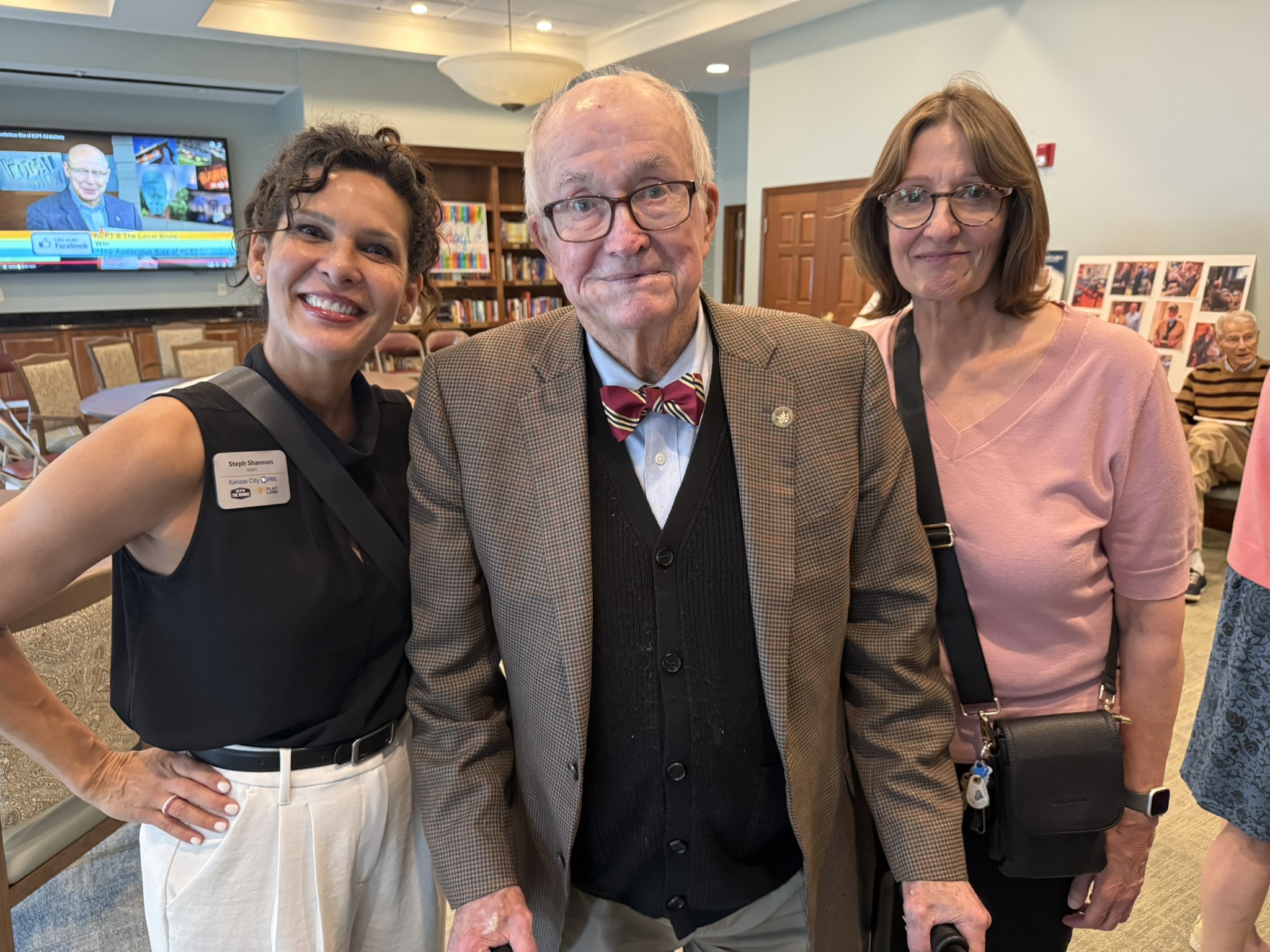 Three people standing indoors, smiling, with bookshelves and a television in the background.
