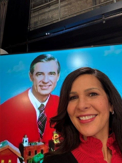 A woman with dark hair smiling in front of a large poster of a man in a red sweater and tie.