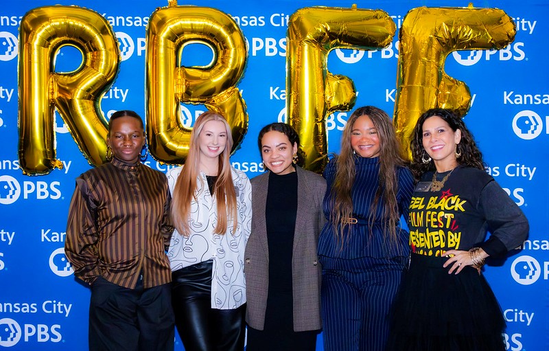 Five people stand in front of a blue Kansas City PBS backdrop, holding large gold balloon letters spelling 'FREE'.