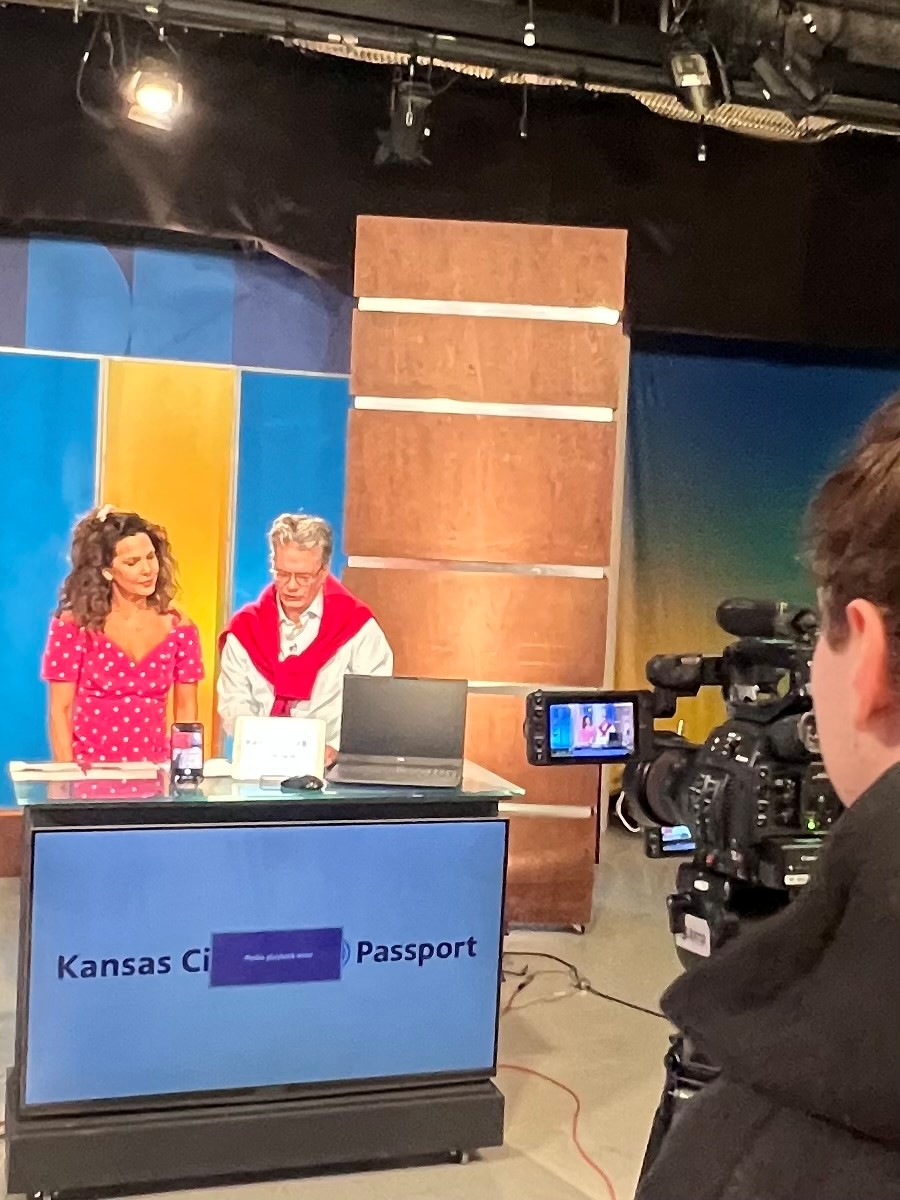 Two women stand behind a desk with a laptop, in a studio with colorful background panels, a camera operator filming them.