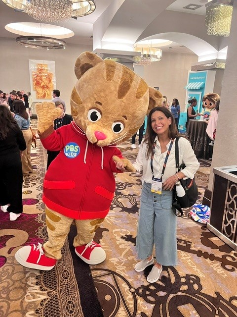 Person in a large cat mascot costume with a red hoodie, standing next to a woman in a white shirt and jeans, in a decorated indoor event space.