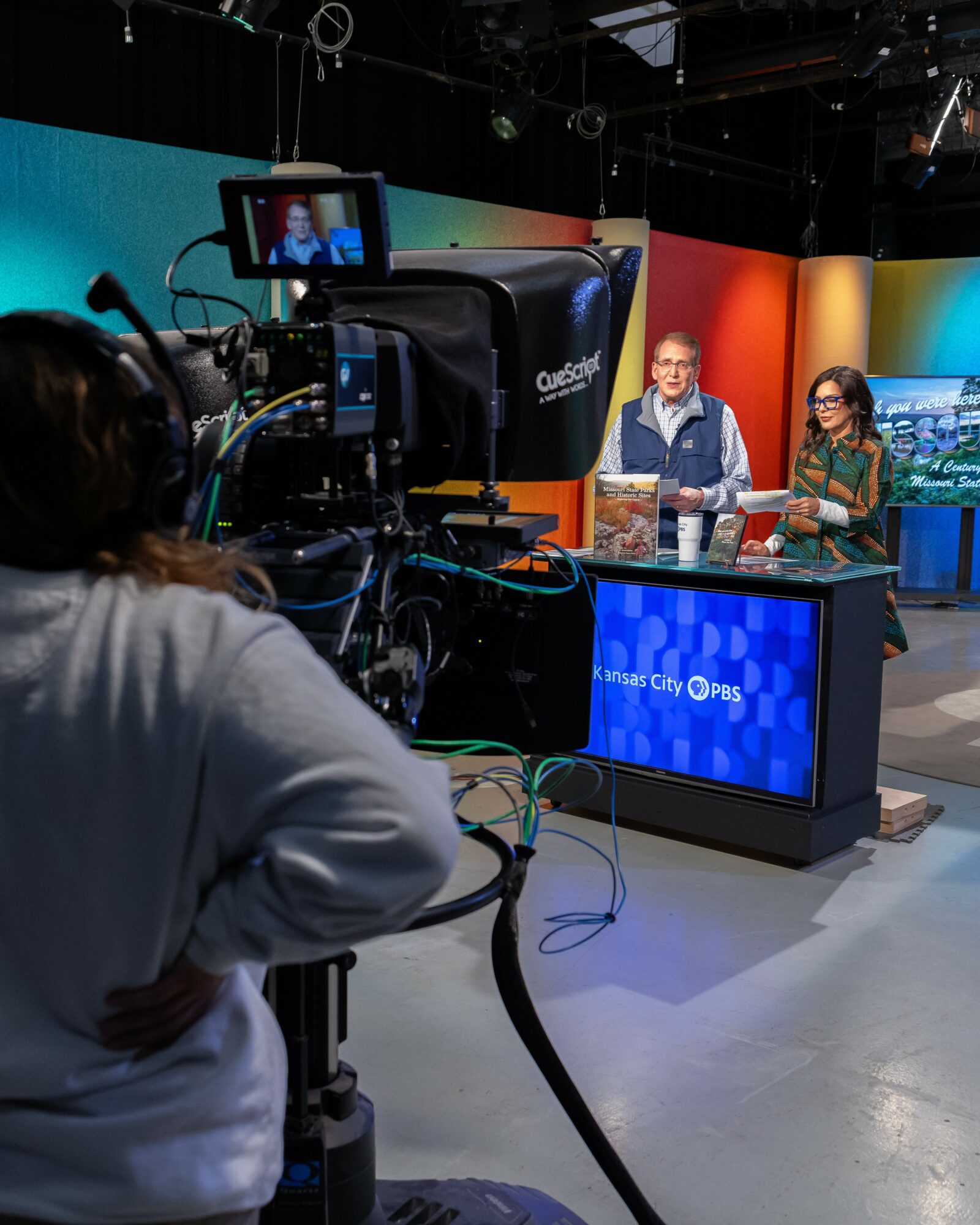 Two people sit at a news desk with a colorful background, being filmed by a camera operator in a studio.