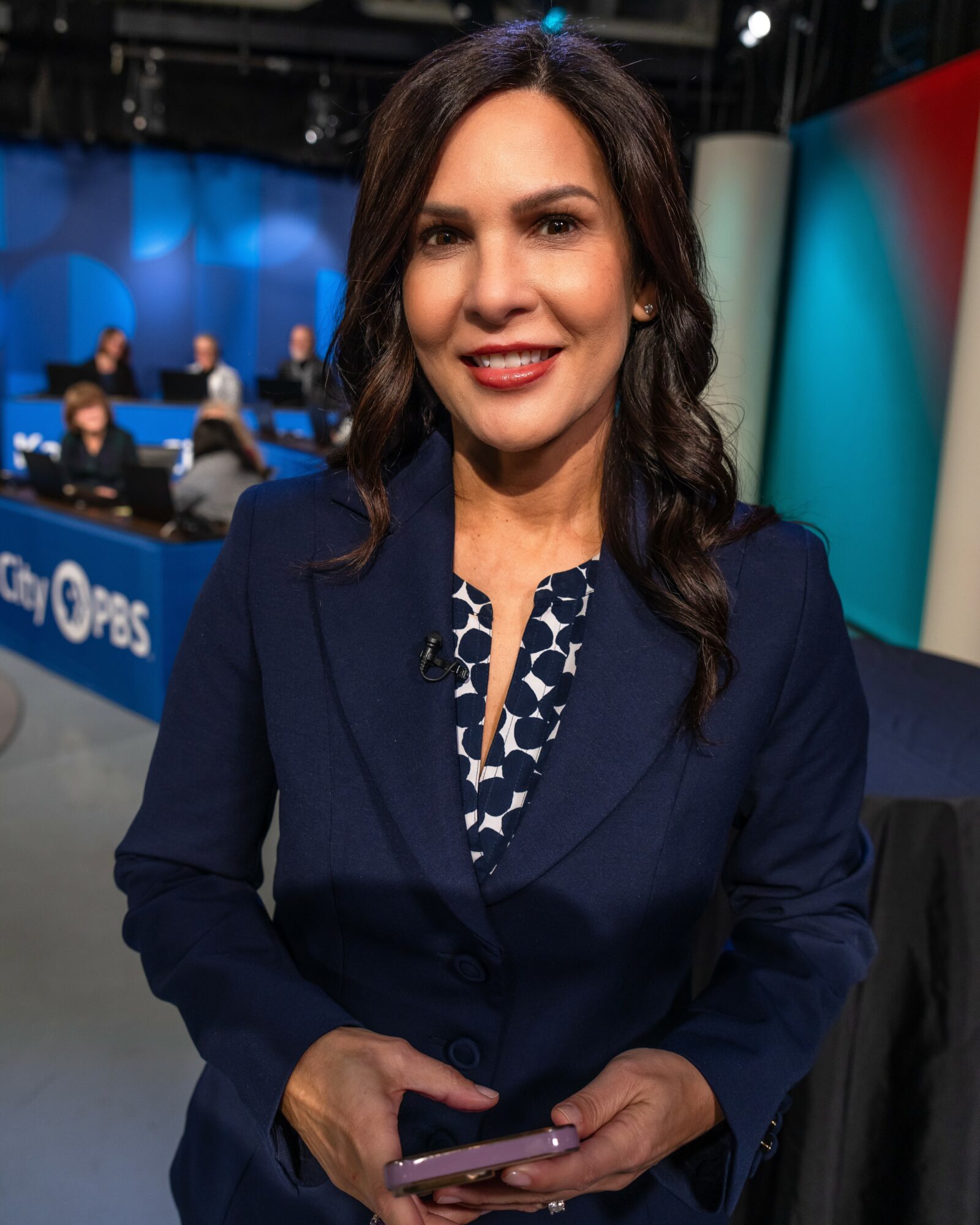 Woman with dark hair in a blue blazer holding a phone, studio background with people and colorful panels.