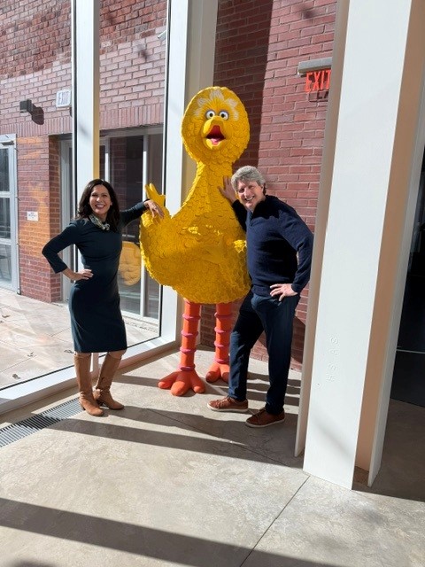 Two people stand beside a large yellow duck sculpture inside a building near windows, with brick walls outside visible.