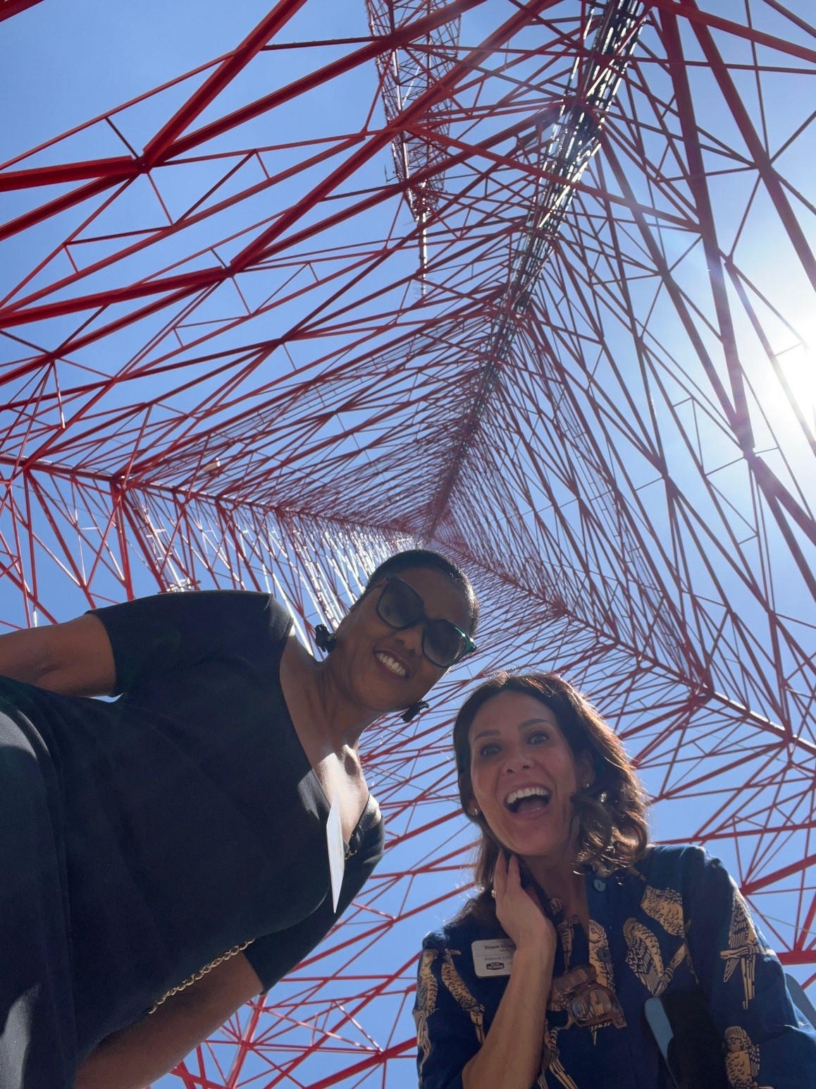 Two women smiling under a large red and white lattice tower against a blue sky.