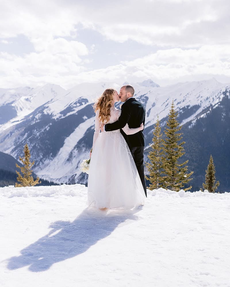 Couple in wedding attire standing on snow-covered ground with mountains and trees in background.