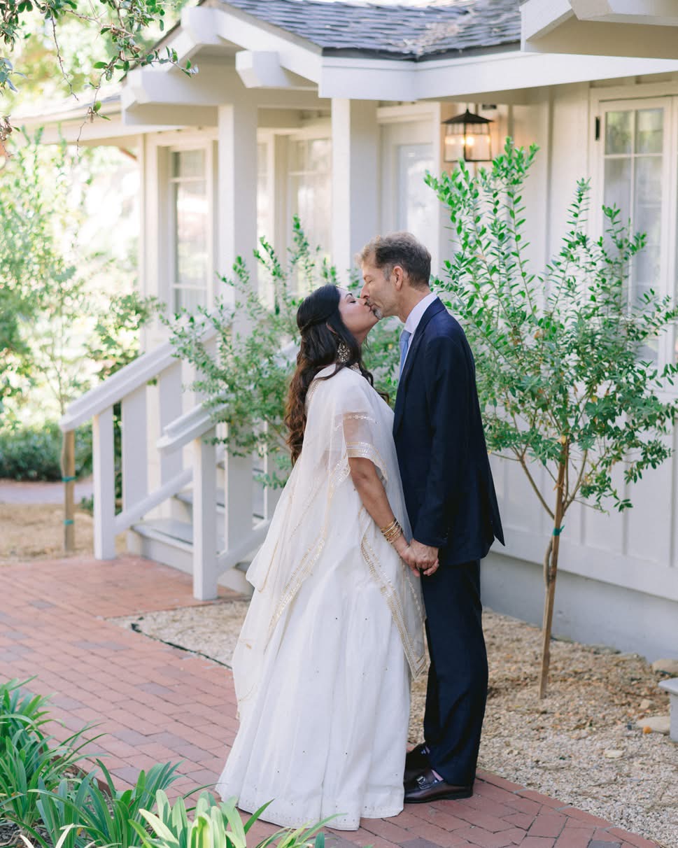A couple in wedding attire holding hands and kissing outside a house with greenery and trees.