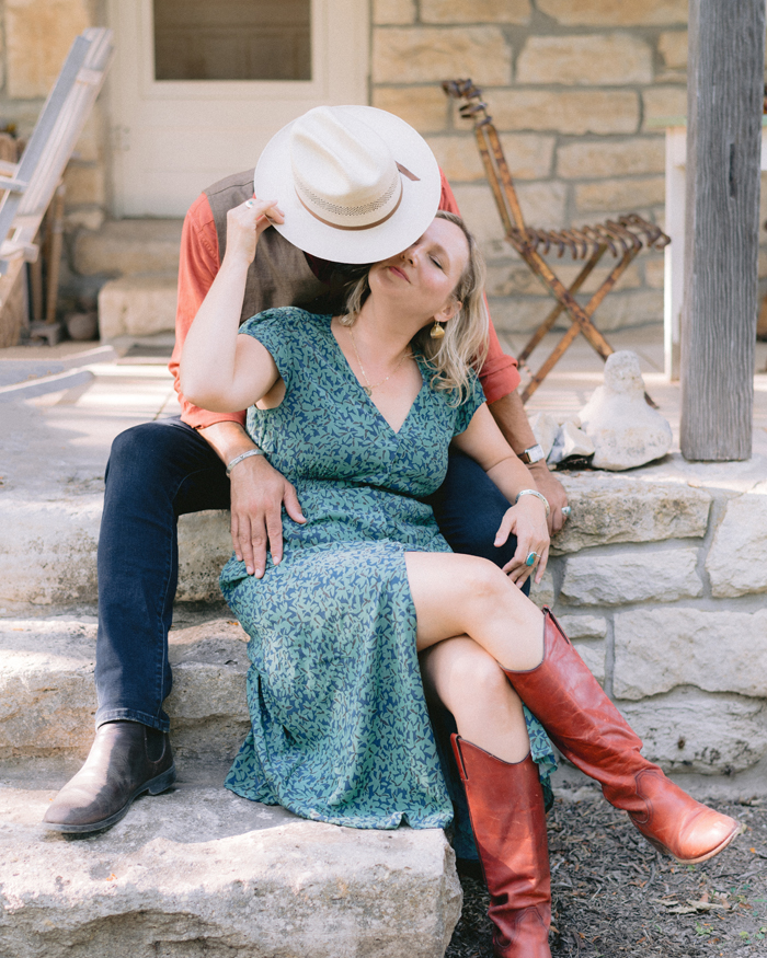 Two women sit on stone steps, one wearing a hat and the other in a blue dress with red boots, kissing.