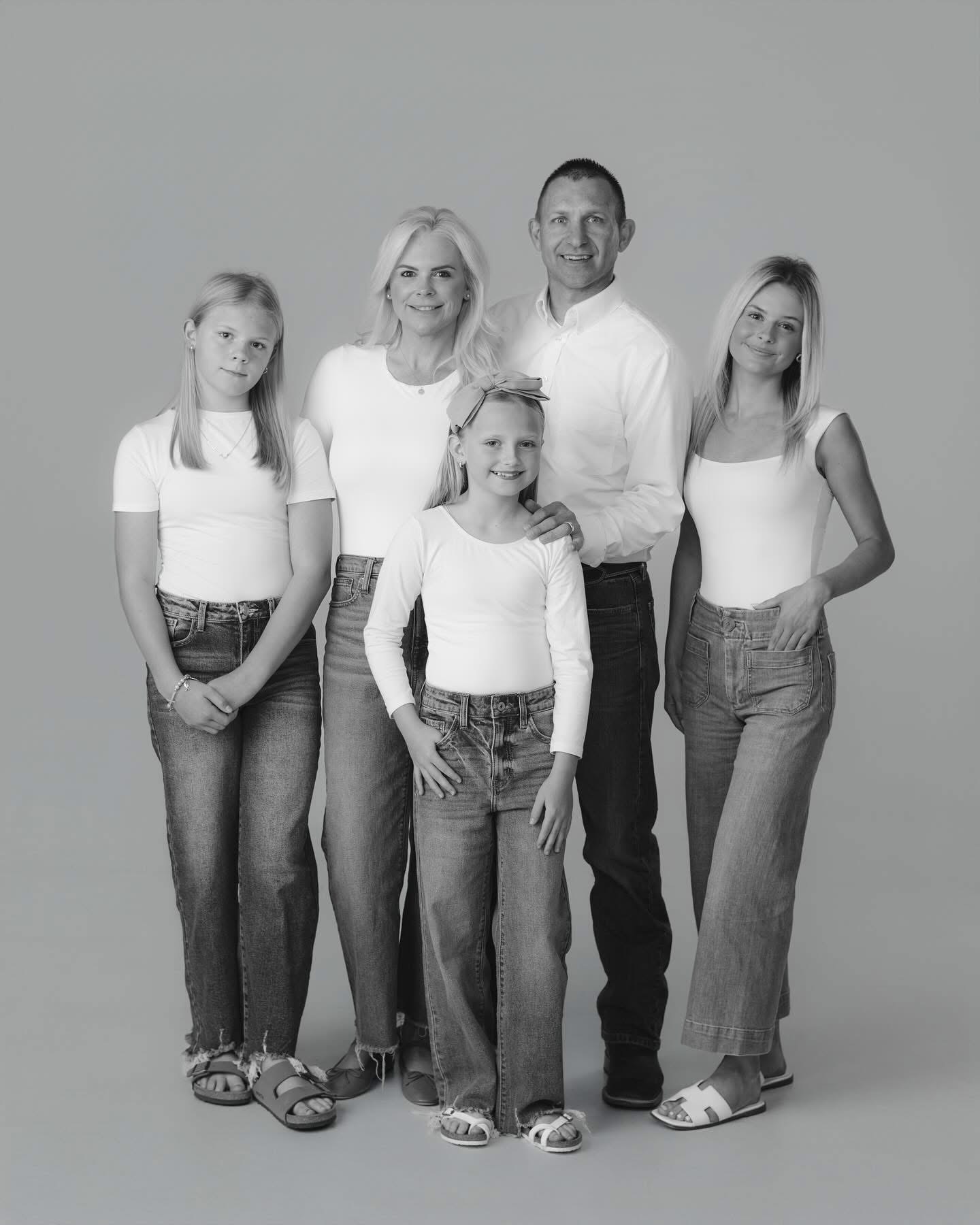 Family of five standing together, all wearing white tops and jeans, smiling, in a studio setting.