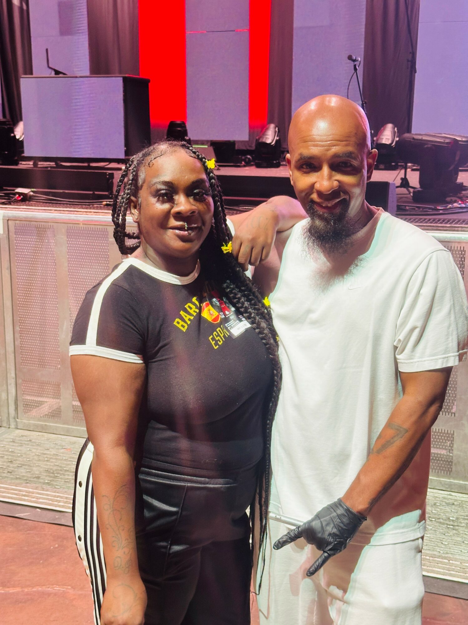 Two people standing together, one with braided hair and the other bald, in a room with stage equipment in the background.