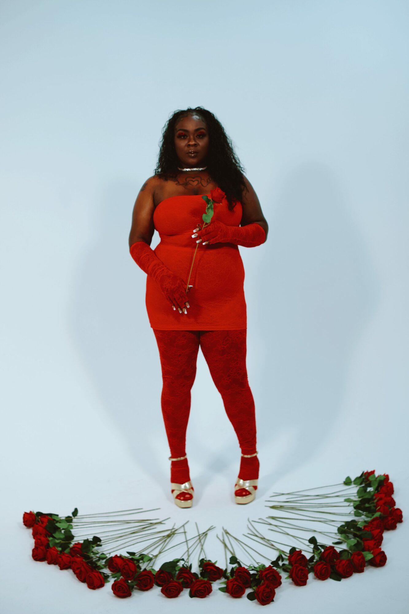 Woman in red dress and gloves stands on a white background with red roses arranged in a semi-circle in front of her.