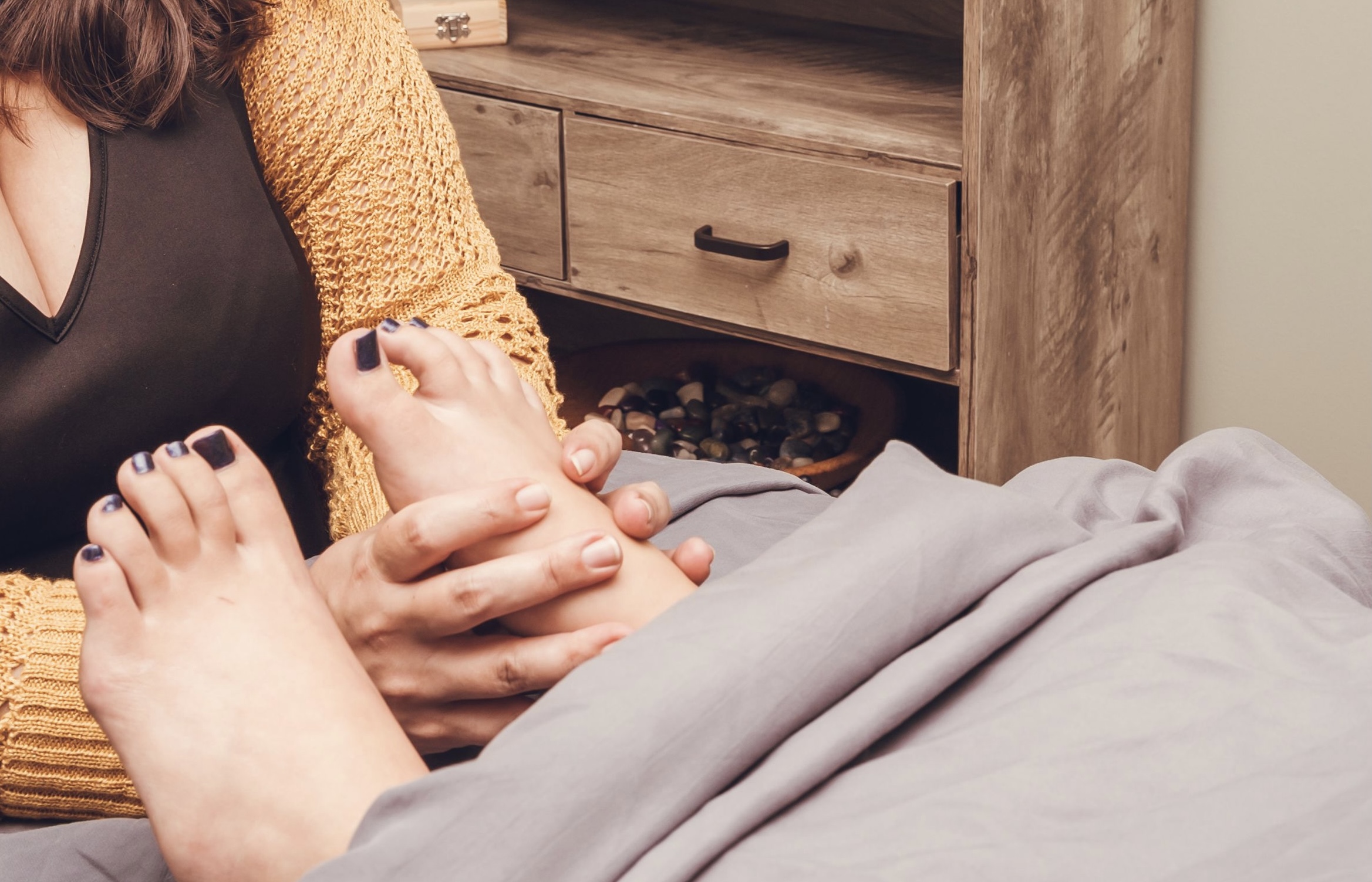 Person holding a baby's foot while lying in bed, with a wooden nightstand in the background.