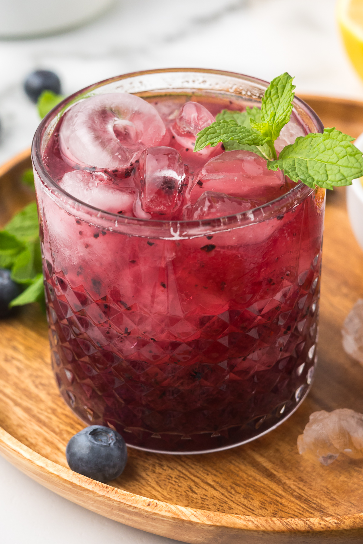 A glass of purple berry drink with ice and mint on a wooden tray, with blueberries nearby.