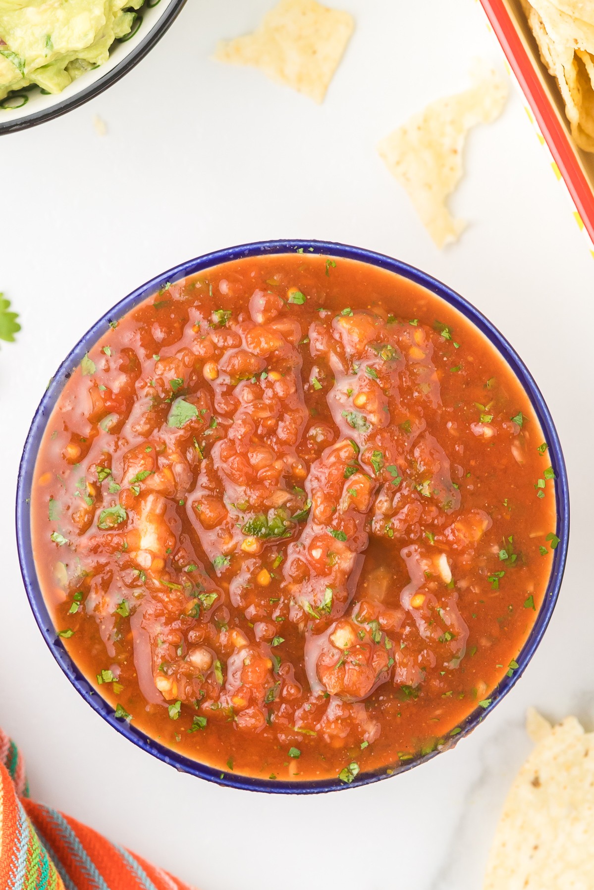 Bowl of spicy tomato-based stew with chunks of meat and green herbs, on a white surface with chips and vegetables nearby.