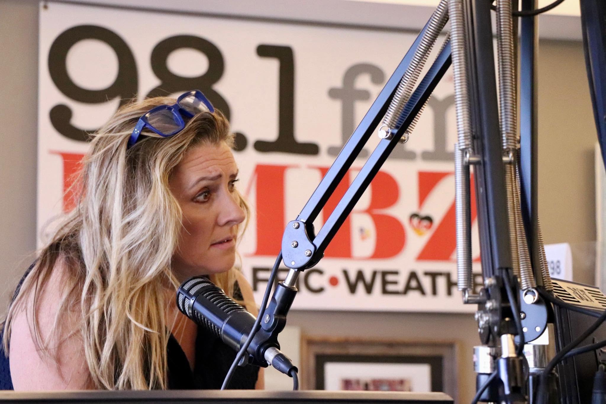 Woman with long blonde hair speaking into a microphone in a radio studio, with a sign in the background.