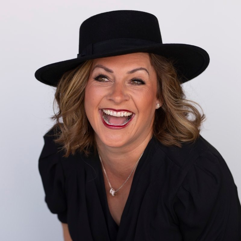 Woman with wavy hair wearing a black hat and black top, smiling broadly, against a plain light background.