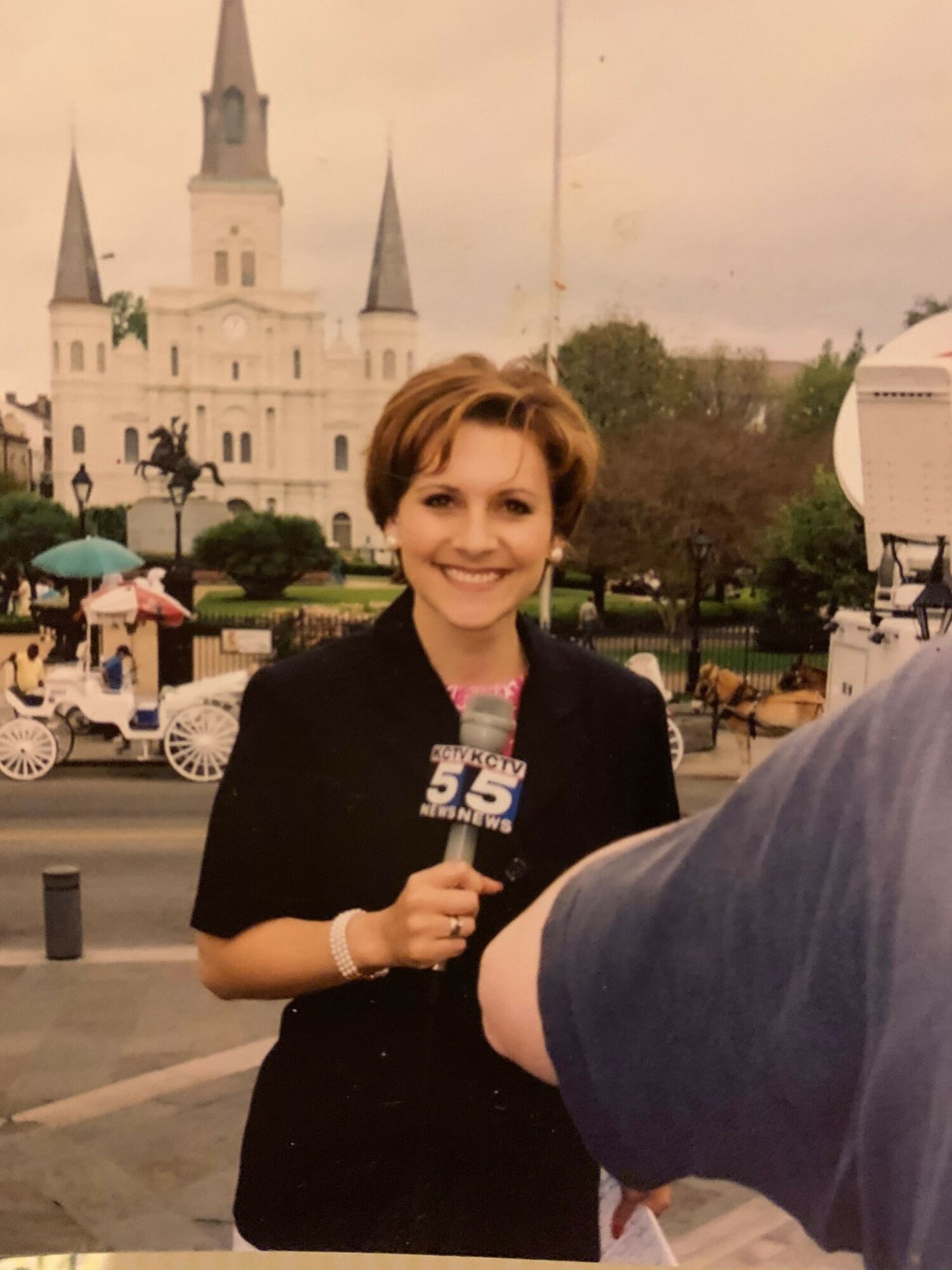 Woman with short hair smiling, holding a drink, standing outdoors near a historic building with a tower, during daytime.