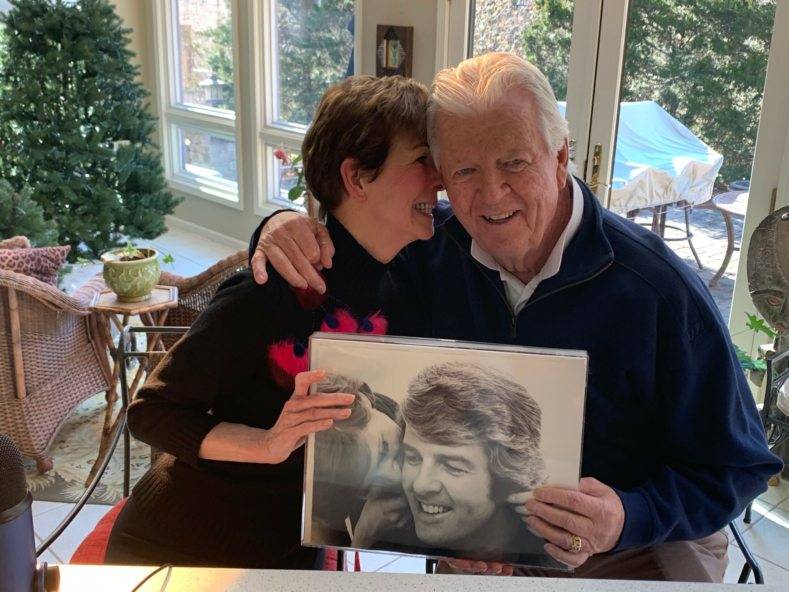 Woman and elderly man smiling, holding a large black-and-white photo of a young man, in a sunlit room with windows and outdoor scenery.