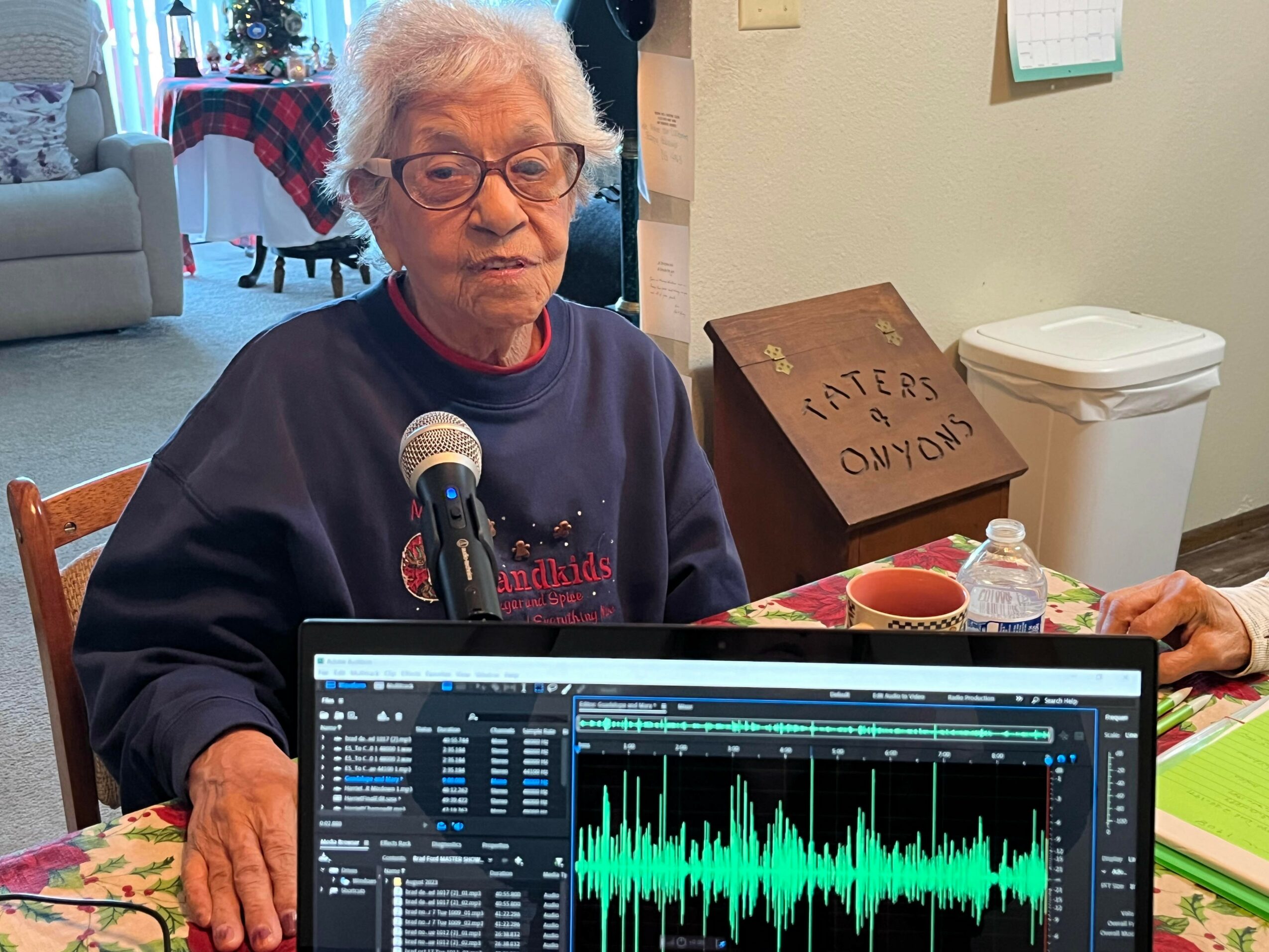 An elderly woman with glasses sits at a table with a microphone, in a room with a couch and a sign that reads 'Haters on my own'.