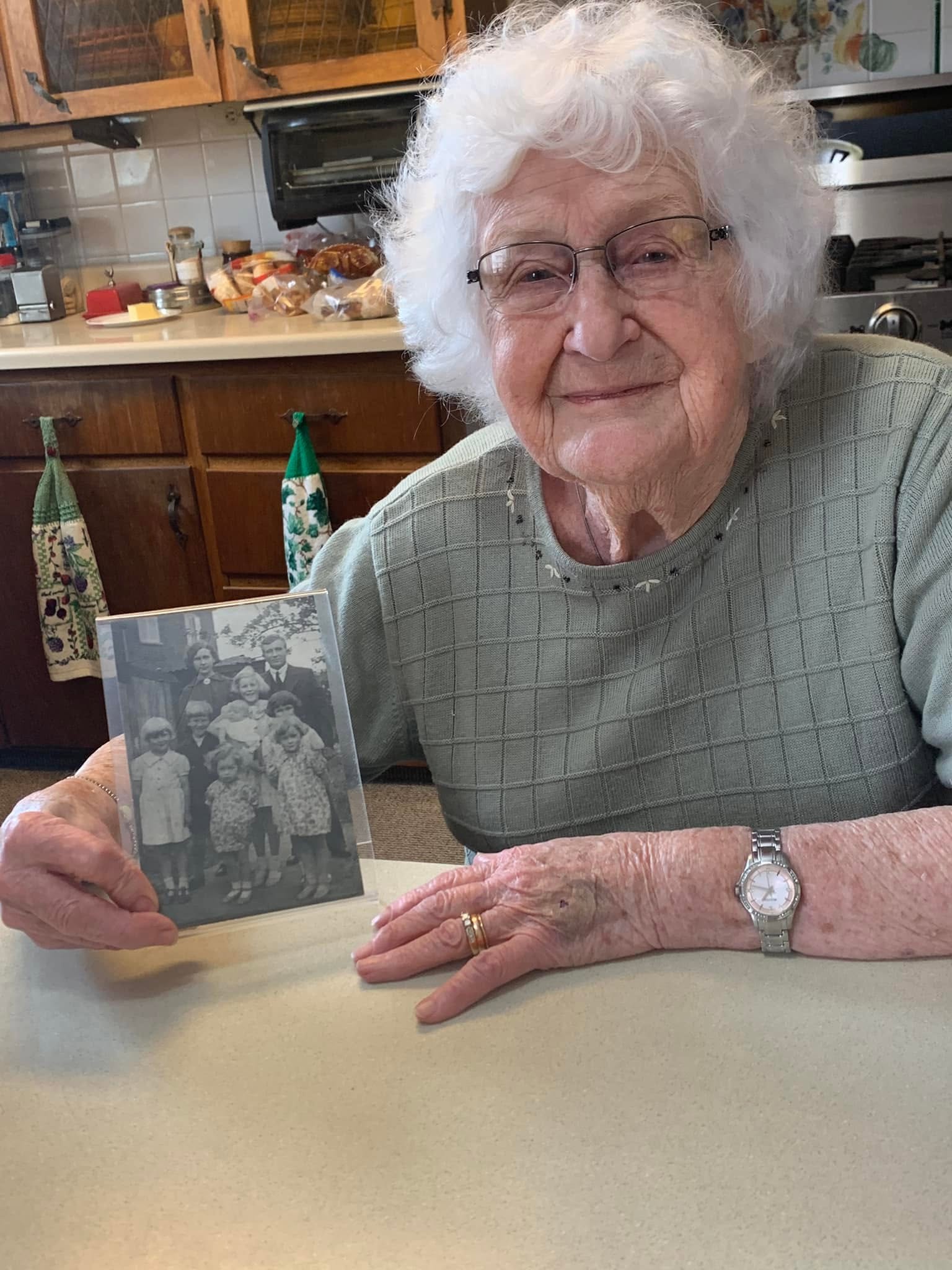 Older woman with white curly hair and glasses holding a black-and-white photo, sitting at a table in a kitchen.