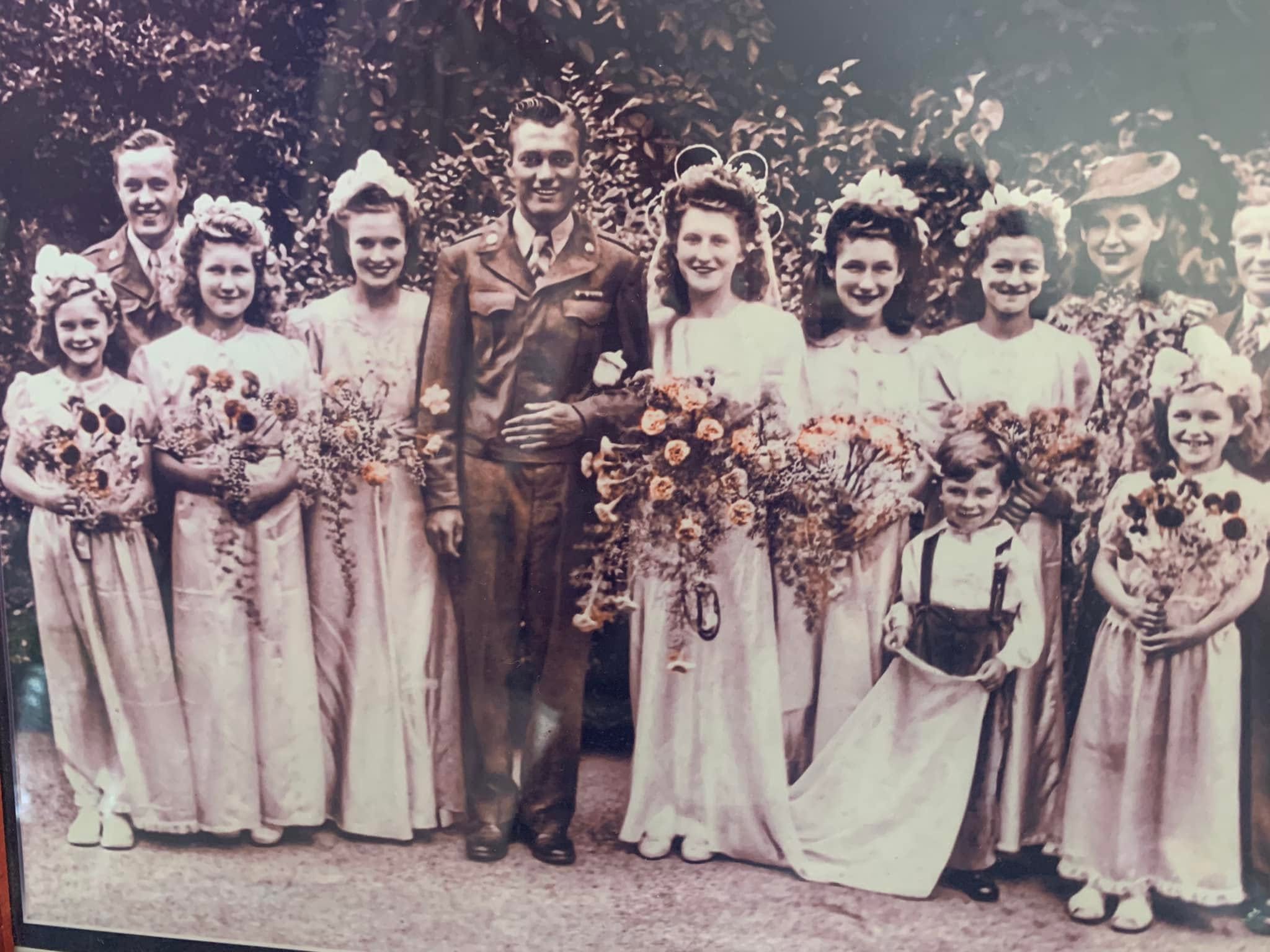 Group of people, mostly women in long dresses, standing outdoors with trees in background, some holding flowers.