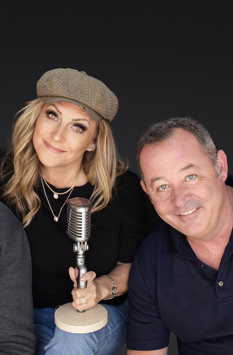 Woman with blonde hair wearing a beige hat and black top, holding a vintage microphone, smiling, man in navy shirt beside her, smiling.
