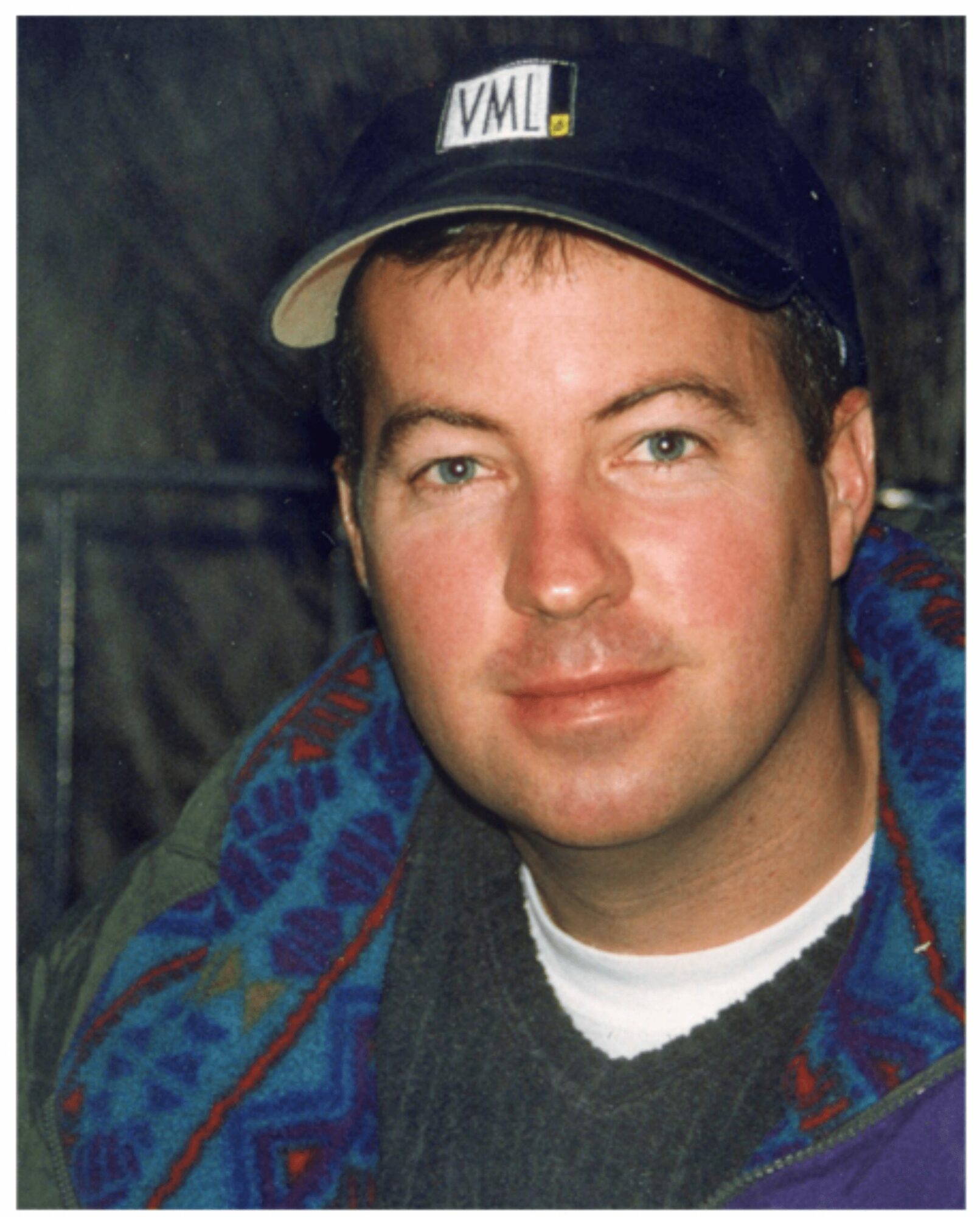Young man wearing a black cap with a logo, smiling, outdoors with trees in the background.