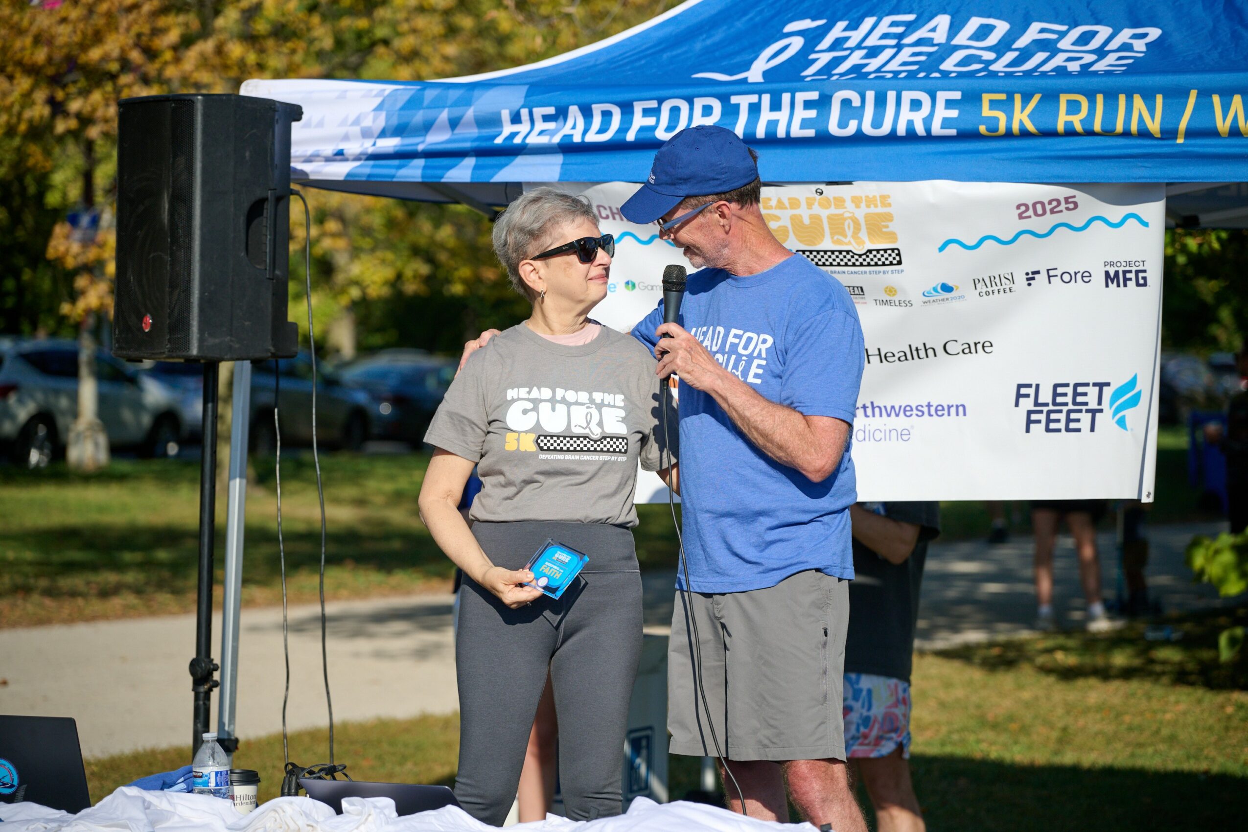 Two people speaking into a microphone outdoors at a charity event, with a blue banner overhead and trees in the background.