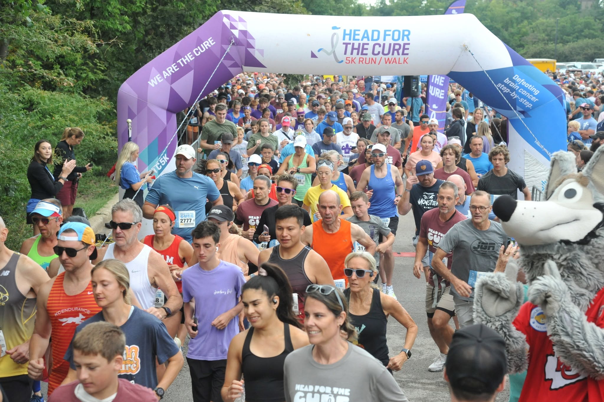 Crowd of people participating in a race under an archway with a mascot on the right side, trees in the background.