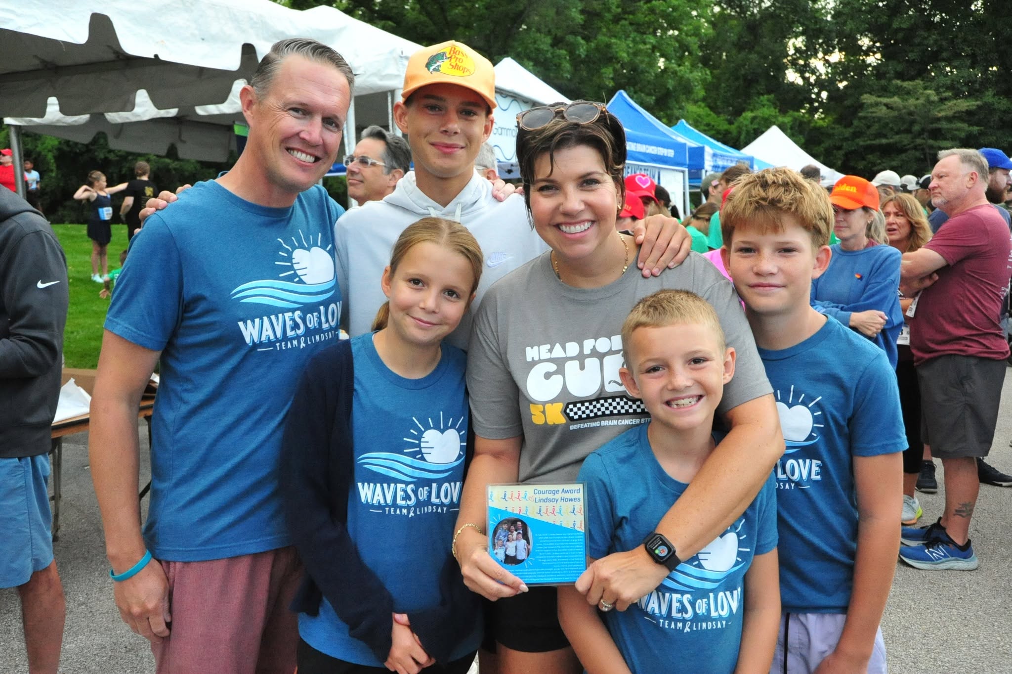 Group of six people smiling outdoors at an event, some wearing matching blue shirts, with tents and trees in background.