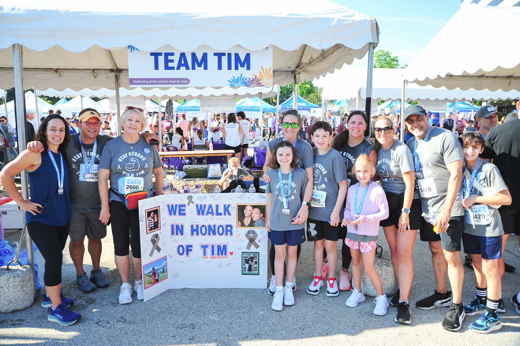 Group of people standing outdoors under a tent, holding a sign that reads 'We walk in honor of Tim,' with a crowd in the background.