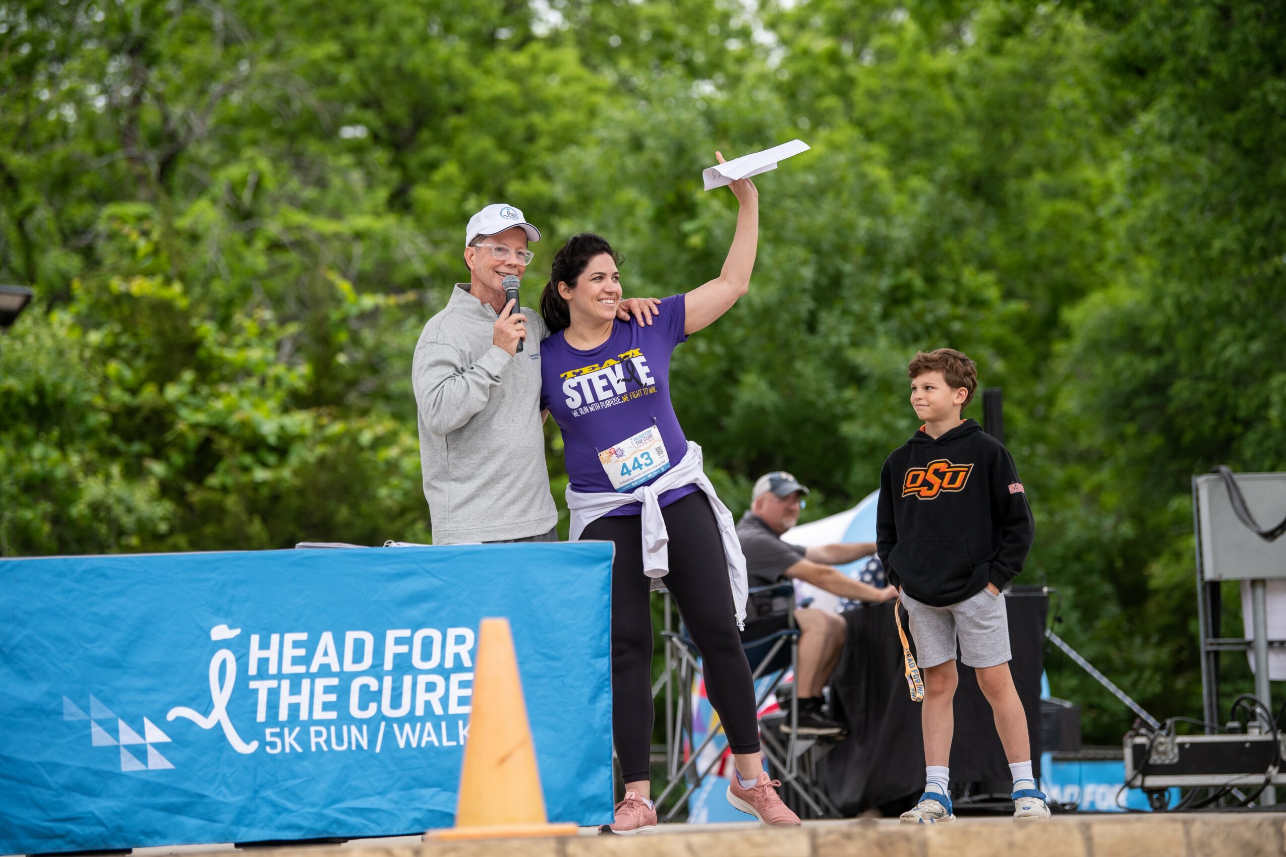 Woman holding a trophy above her head, smiling, with two men and trees in the background at a race event.