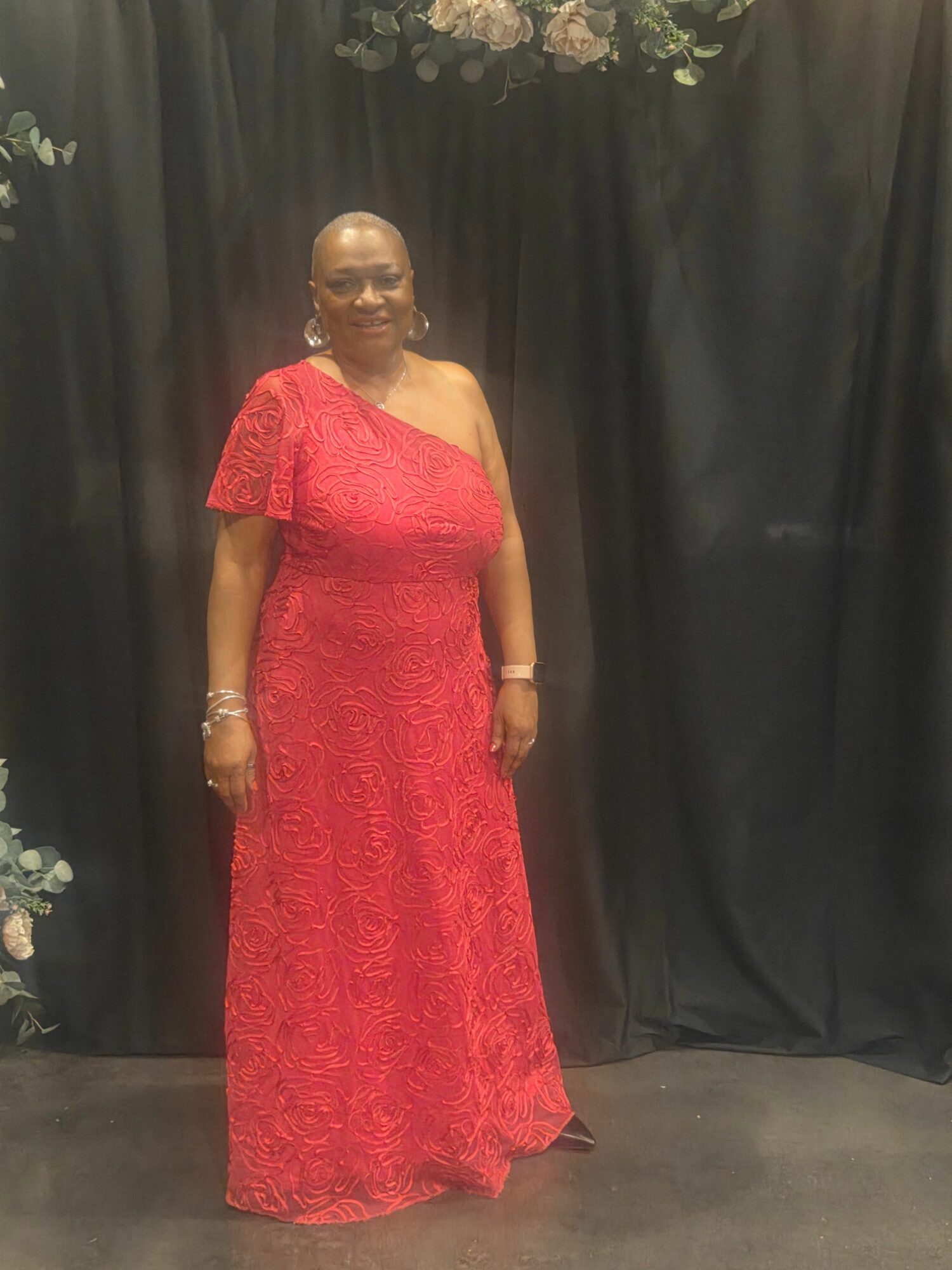 Woman in a red dress standing against a black curtain backdrop, with floral decorations on the side.