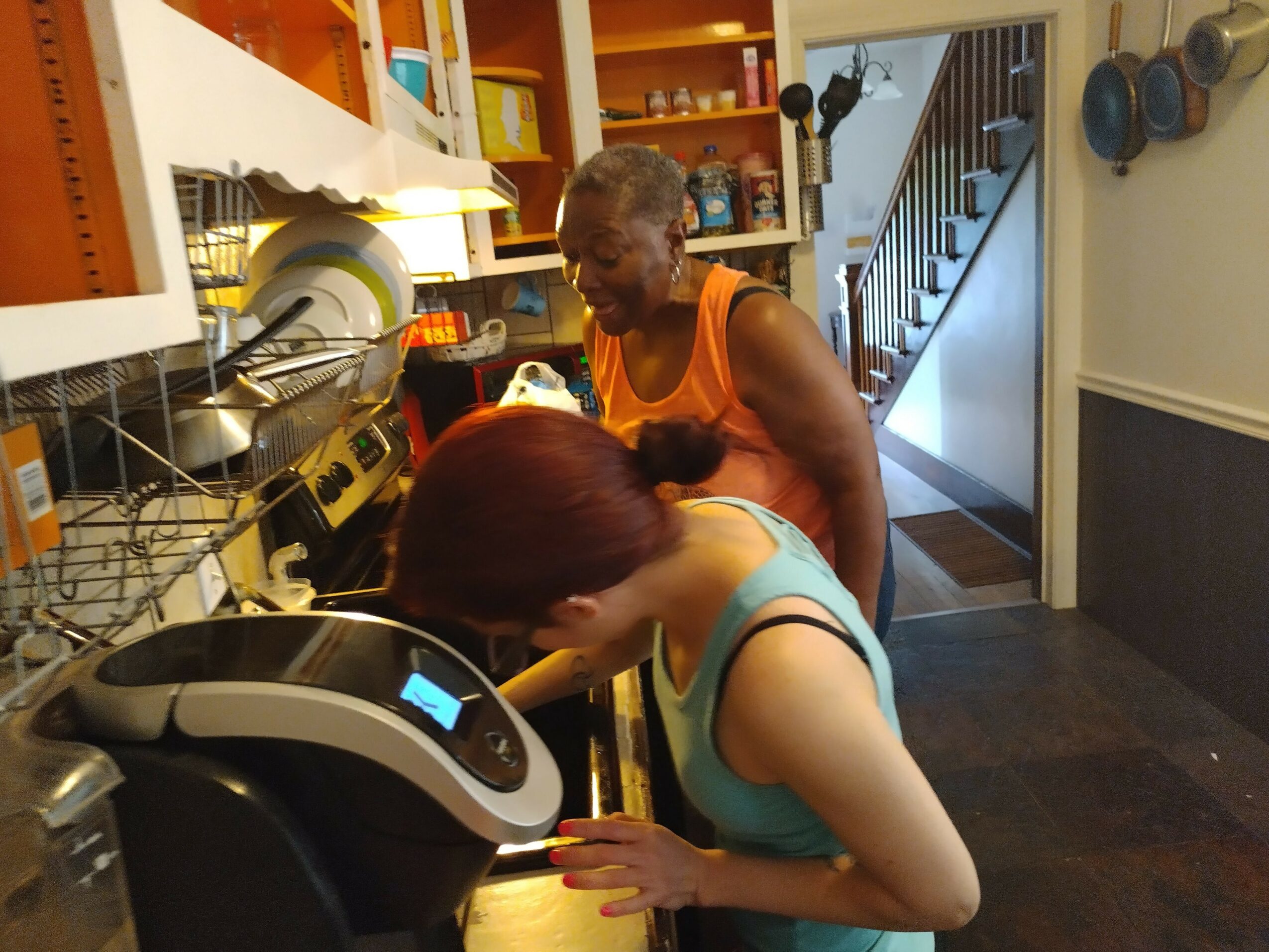 Two women in a kitchen, one leaning over a counter, the other standing behind her, with shelves and a staircase in the background.