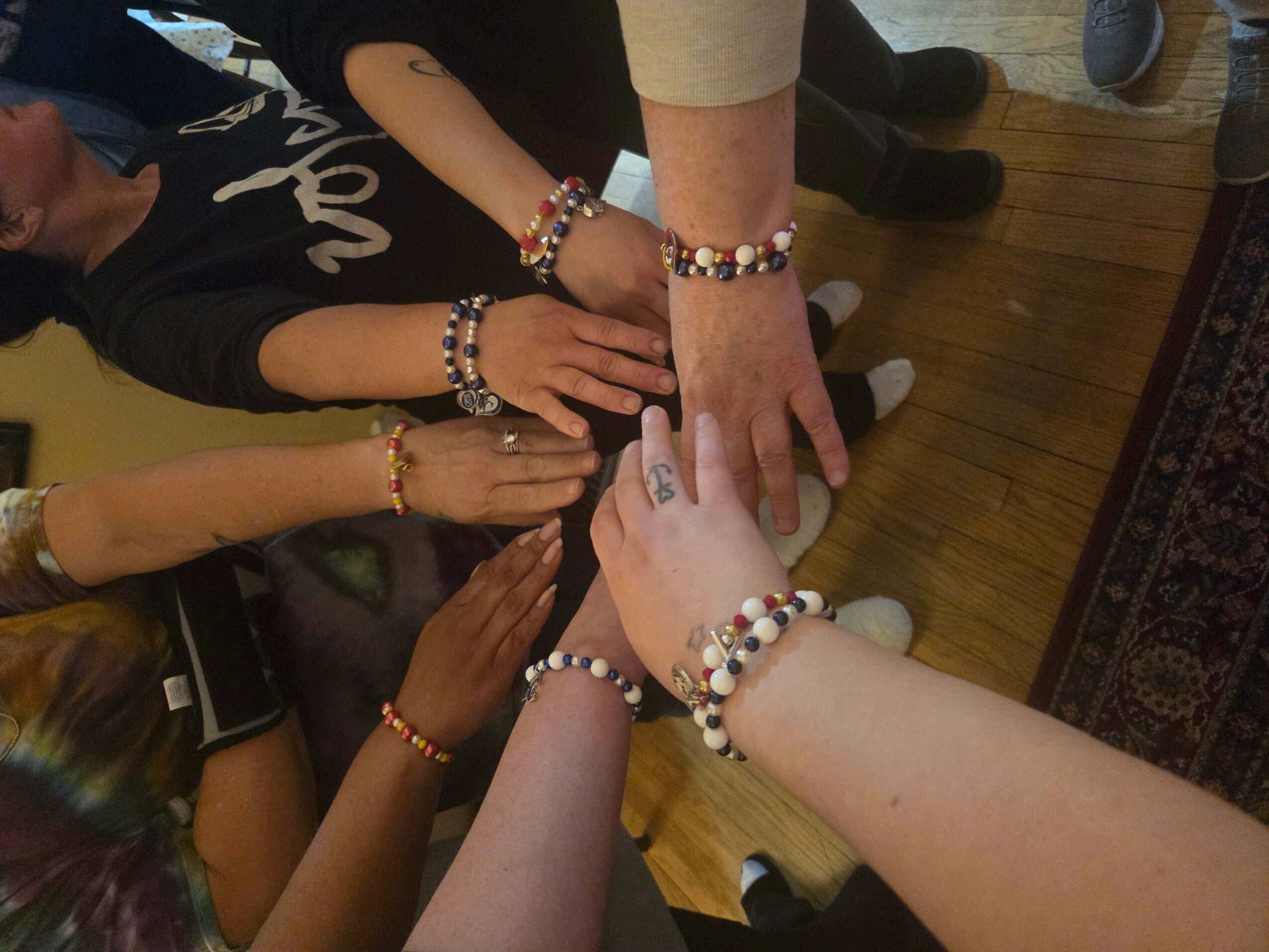 Group of people with hands stacked together, wearing colorful beaded bracelets, on a wooden floor.
