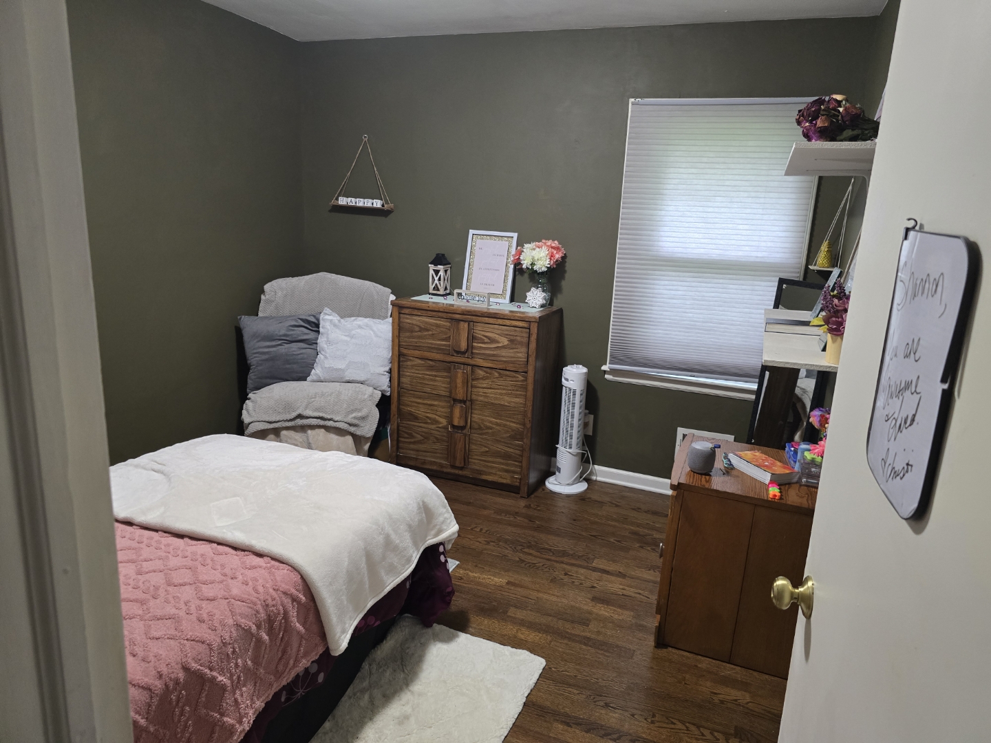 Bedroom with bed, chair, dresser, window, and shelves, decorated with flowers and personal items.