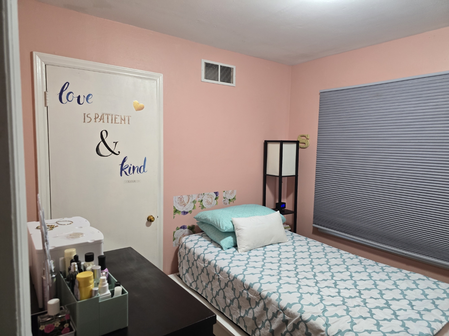 Pink bedroom with a bed, window with closed blinds, and wall decorations, including a door with words and a shelf.