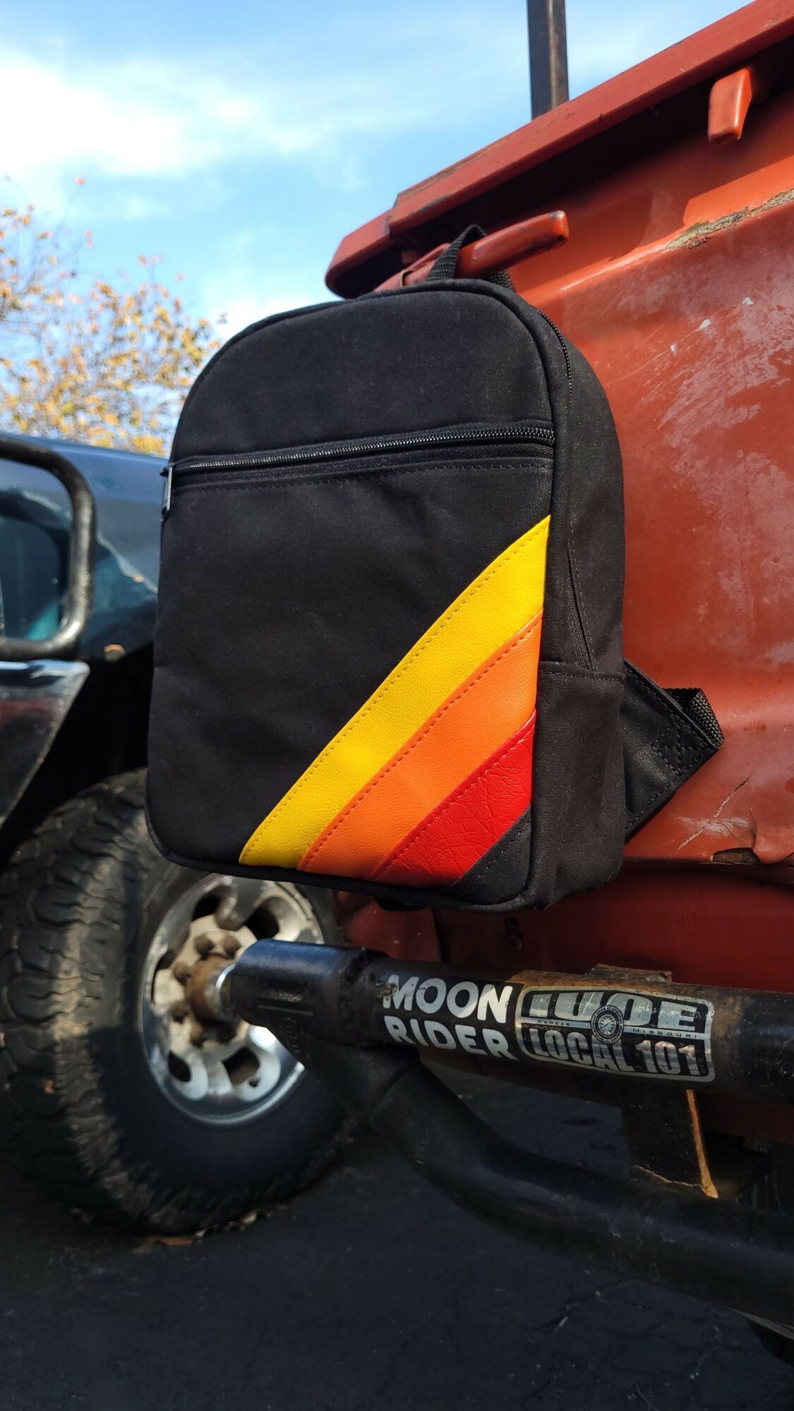 Black backpack with yellow and red diagonal stripes hanging on a red surface outdoors.