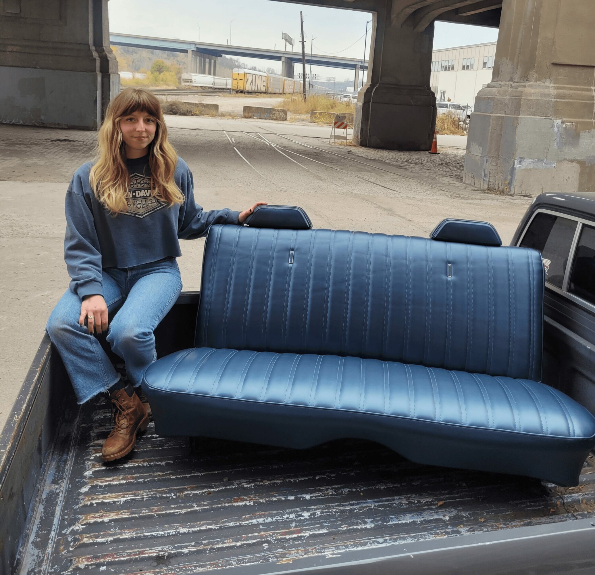 Woman sitting on the edge of a blue vintage sofa in the back of a truck, under a bridge, outdoors.
