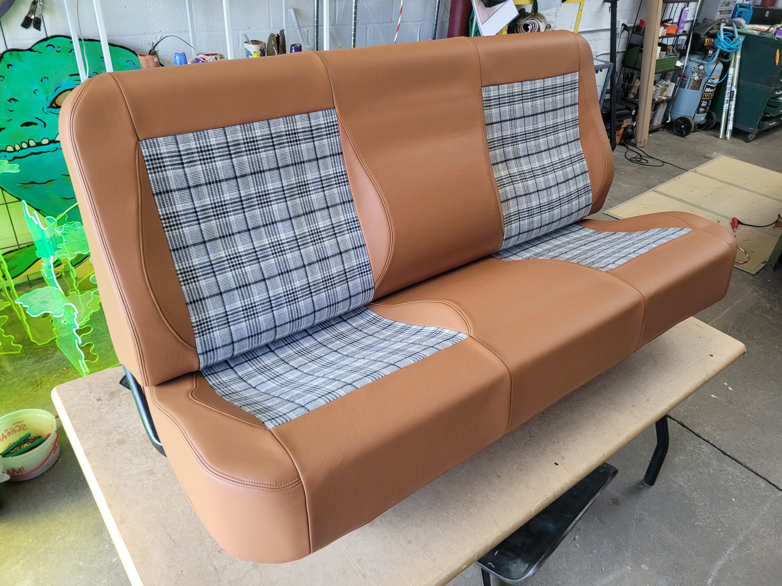 A brown upholstered bench with plaid fabric sections on a beige table in a workshop setting.
