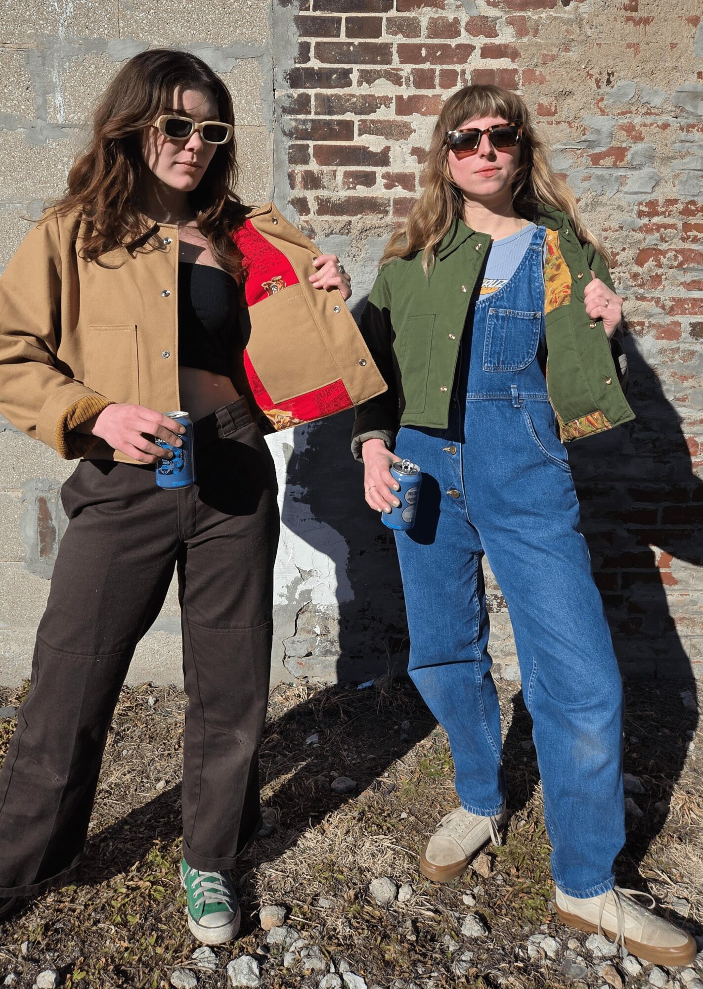 Two women with sunglasses standing outdoors against a brick wall, holding drinks and bags, dressed casually.