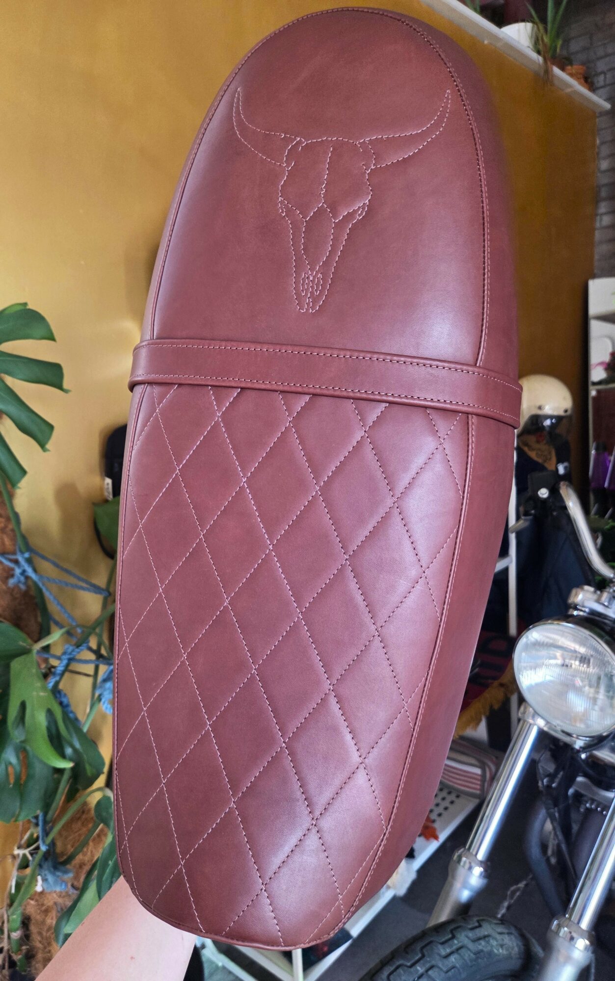 Upright motorcycle seat with quilted pattern and skull logo, surrounded by plants and motorcycle parts.