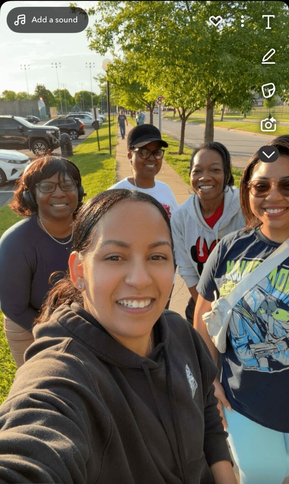 Group of five people smiling outdoors with trees and parked cars in background.