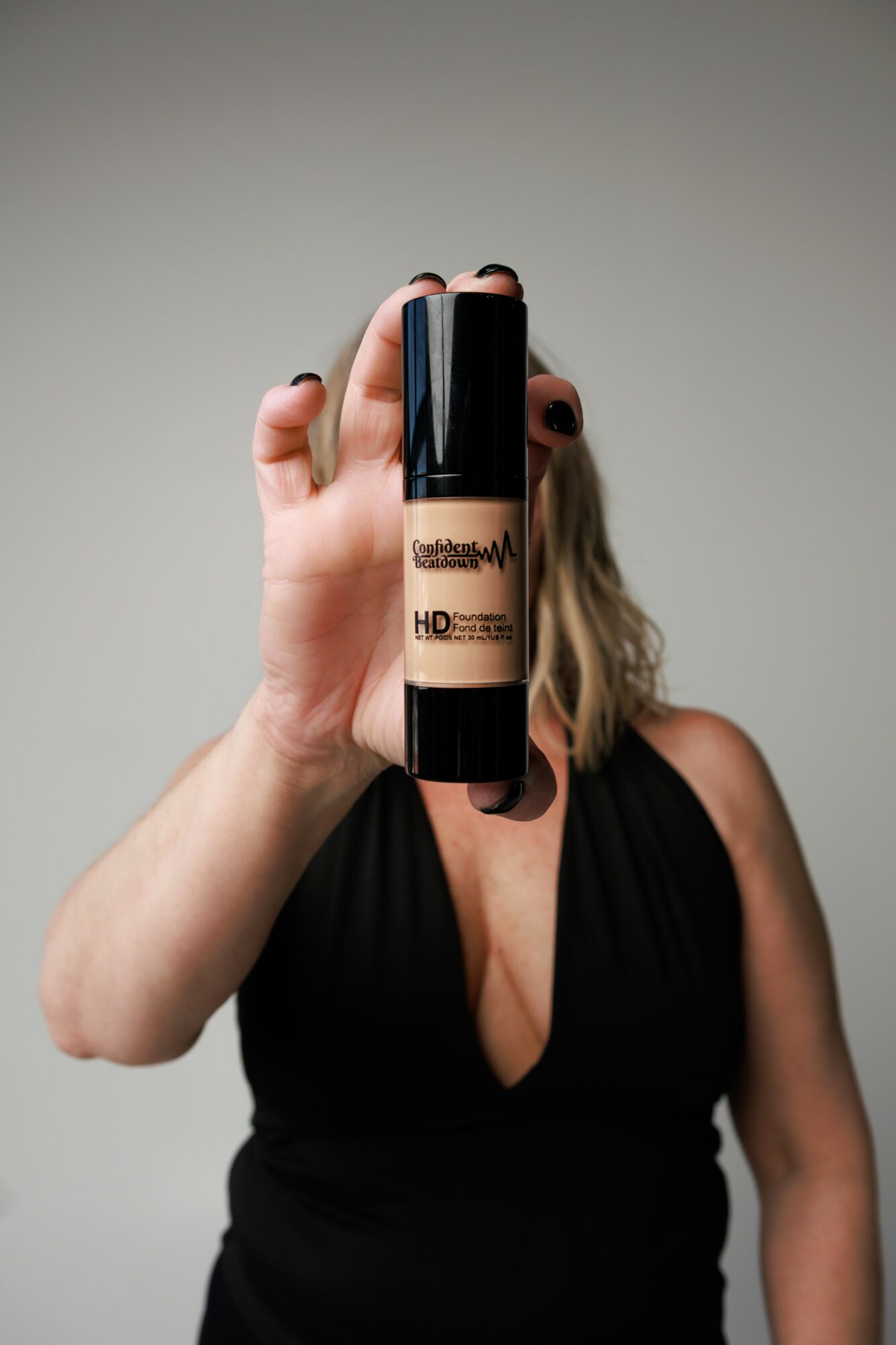 Person holding a bottle of foundation makeup in front of face, wearing a black top, against a plain background.