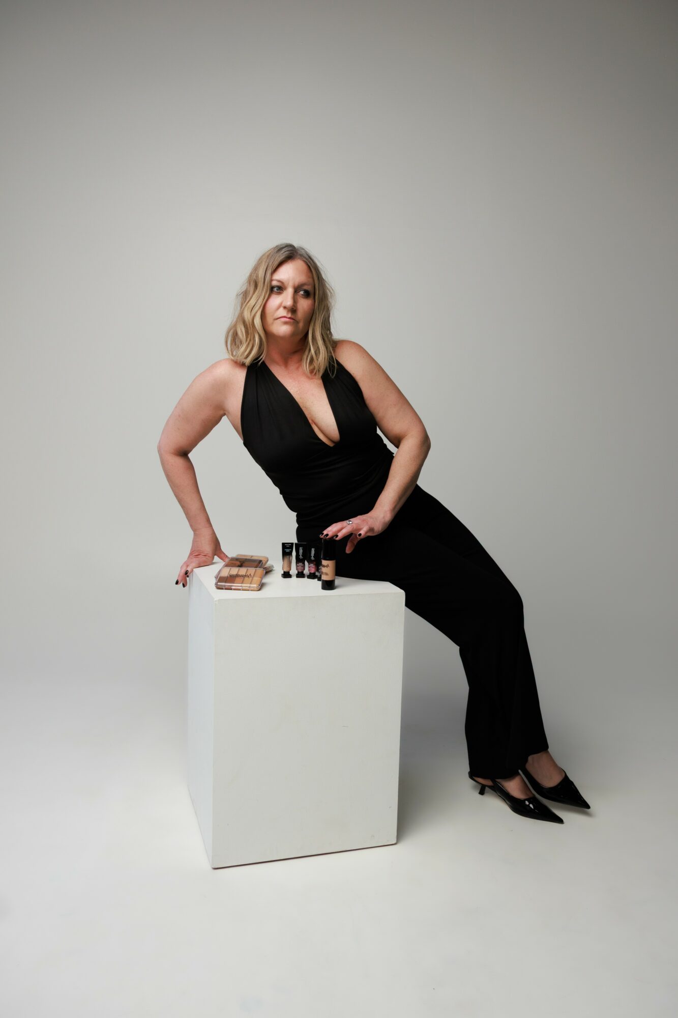 Woman in black dress sitting on a white block with makeup products and brushes, neutral background.