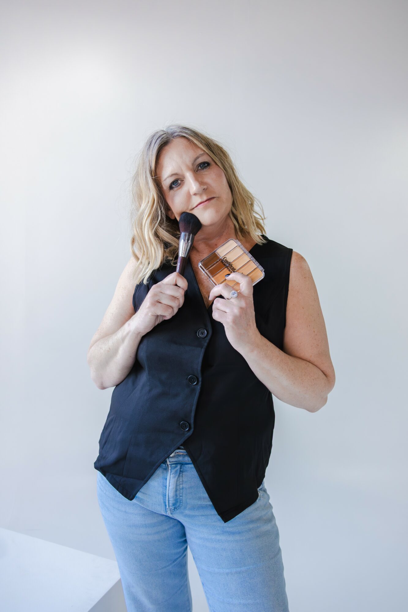 Woman with blonde hair holding a microphone and a small wooden object, standing against a plain light background.