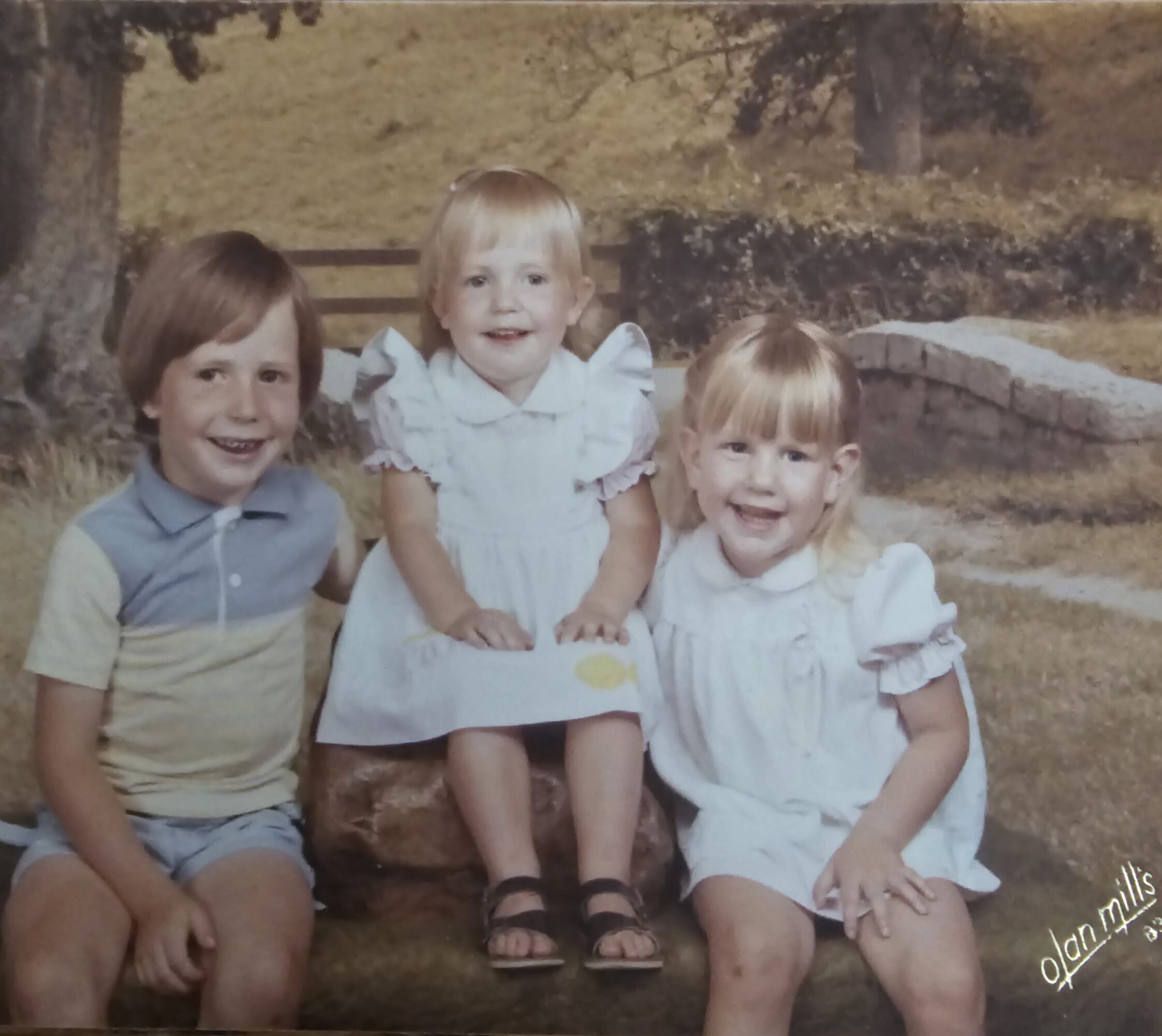 Three children sitting outdoors on a log, smiling, with trees and grass in the background.