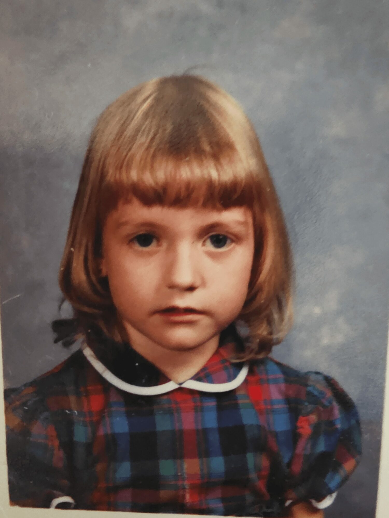 Young girl with shoulder-length light brown hair and bangs, wearing a plaid dress with a white collar, looking at the camera.