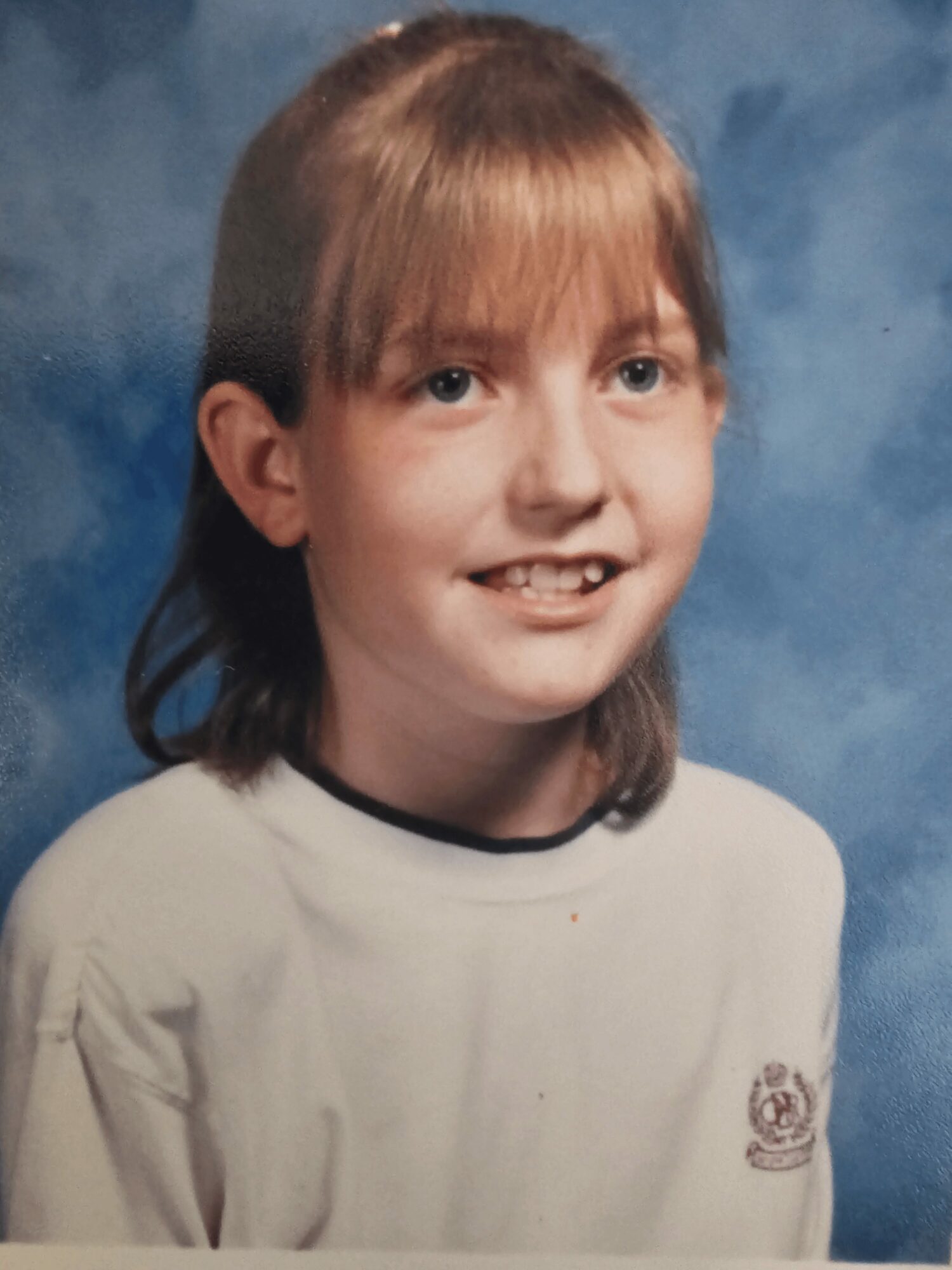 Young girl with light brown hair and bangs, smiling, wearing a white shirt with a small emblem, against a blue background.