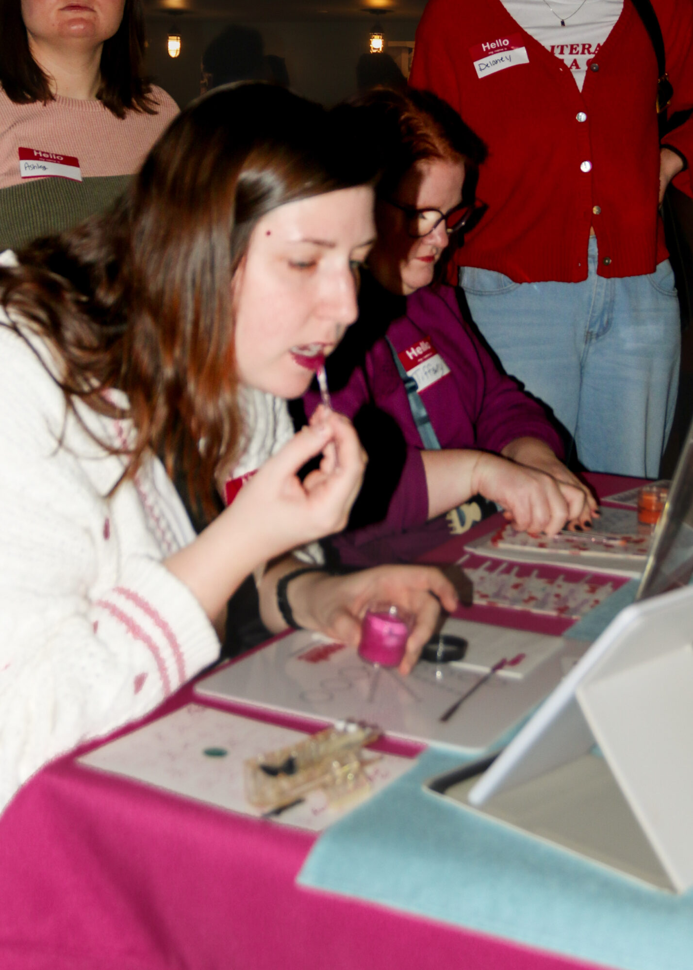 Young woman with long brown hair using a laptop, surrounded by others, with makeup and nail polish on the table.